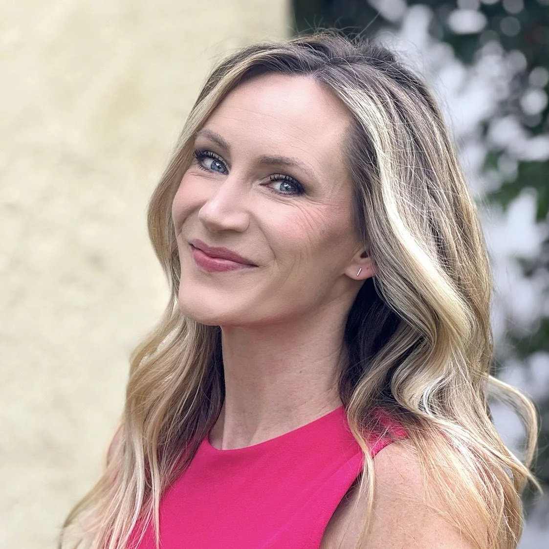 A woman with wavy blonde hair and blue eyes, smiling and wearing a sleeveless pink top, standing outdoors with blurred greenery in the background.