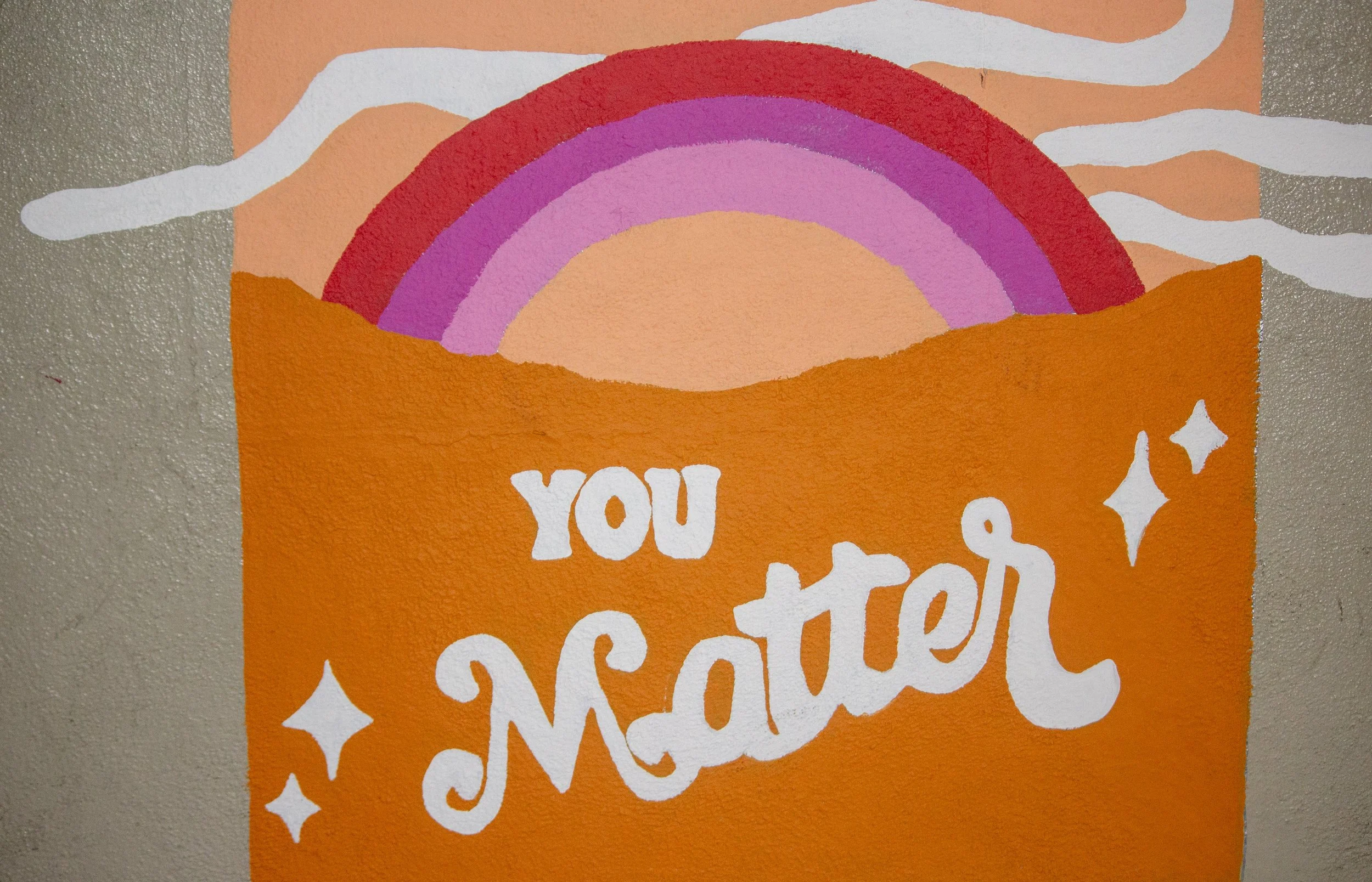Colorful mural featuring a rainbow with pink and purple bands, white clouds, an orange background with the white words "you matter" and star shapes.