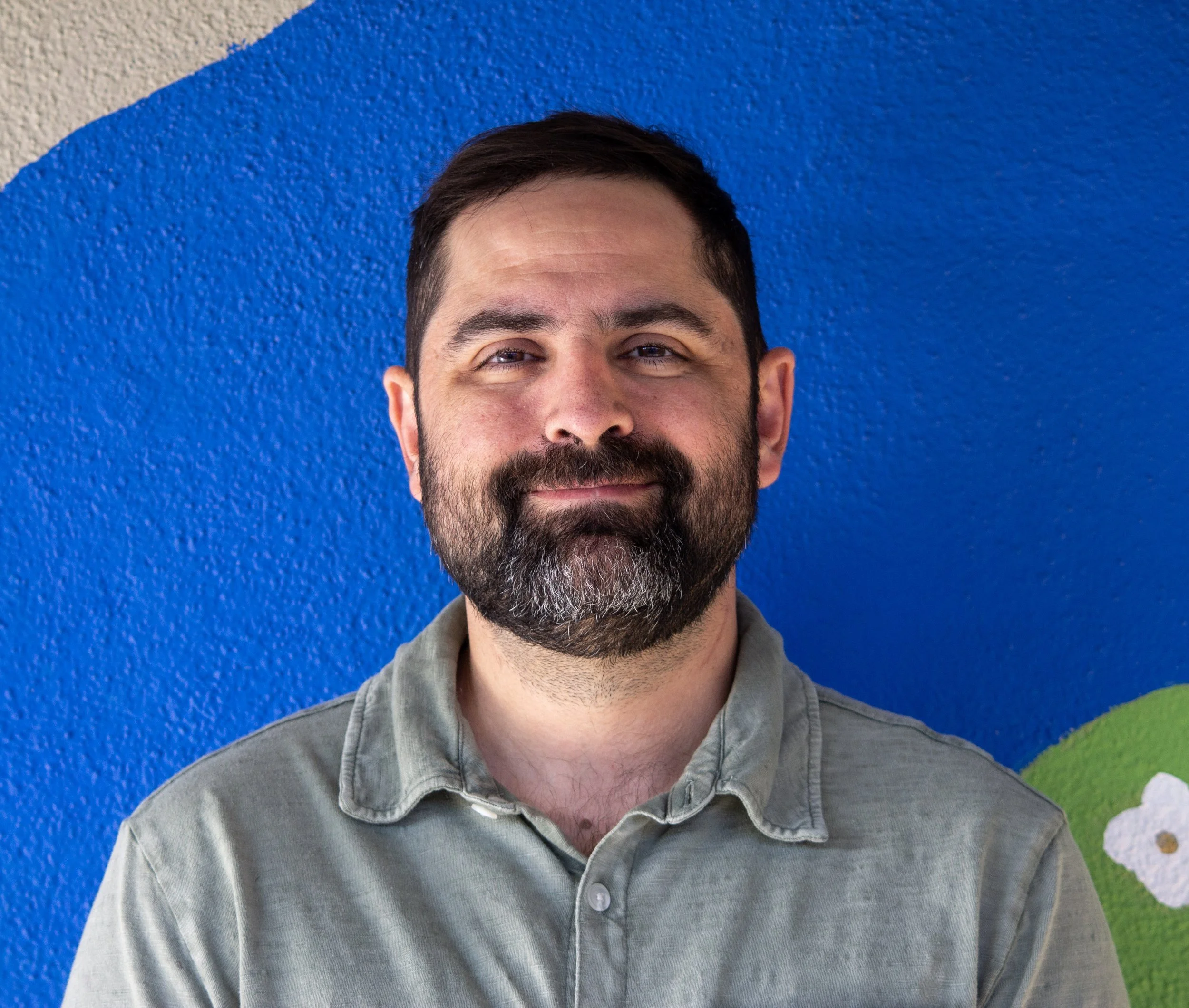A man with a beard and mustache, smiling, wearing a tuxedo and bow tie, against a blue background.