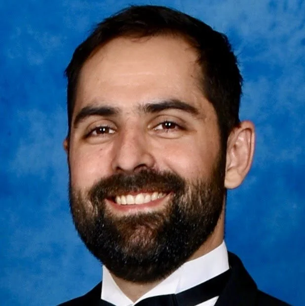 A man with a beard and mustache, smiling, wearing a tuxedo and bow tie, against a blue background.