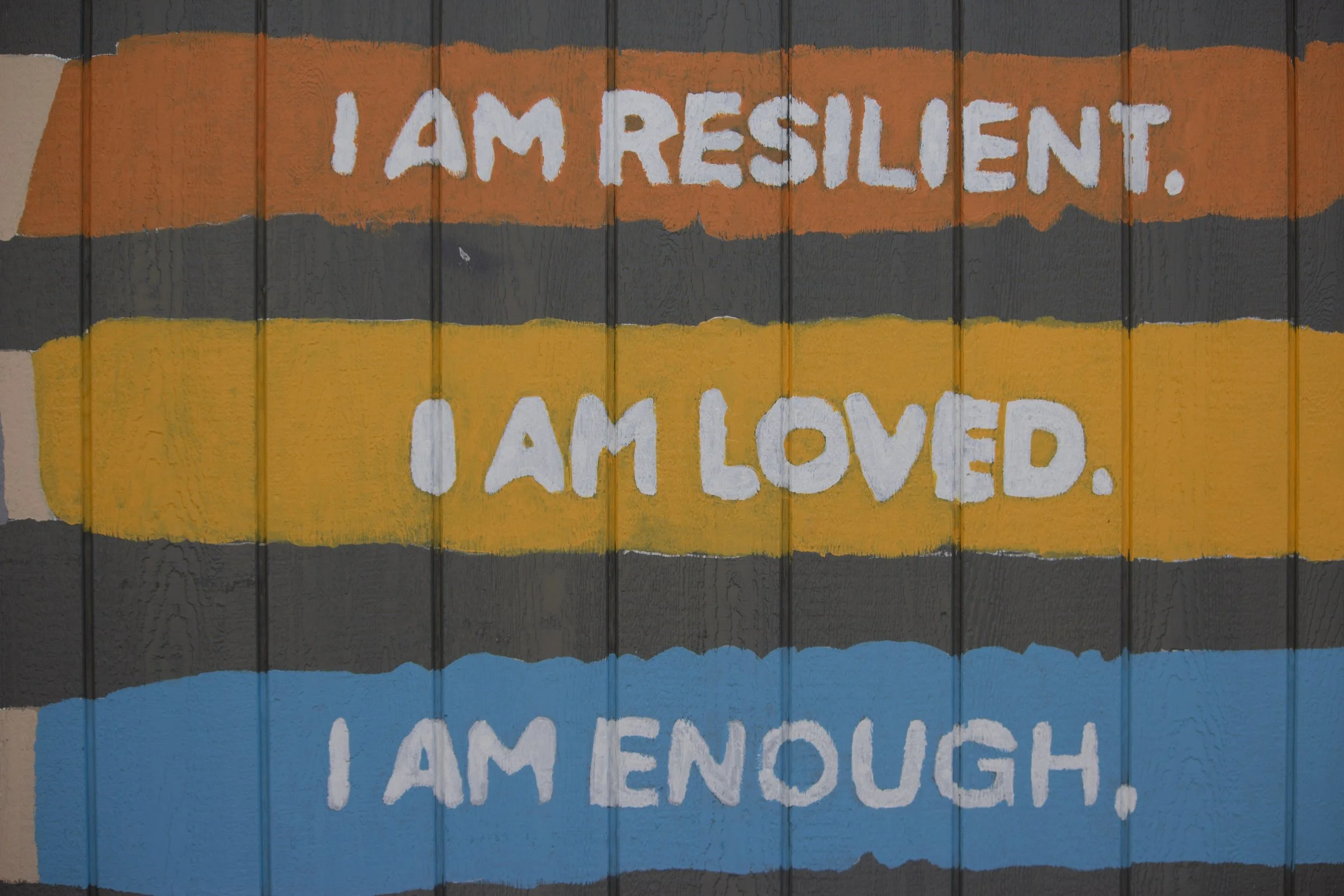 Colorful mural with horizontal stripes in orange, yellow, black, and blue, featuring the white hand-painted message: 'I am resilient. I am loved. I am enough.'