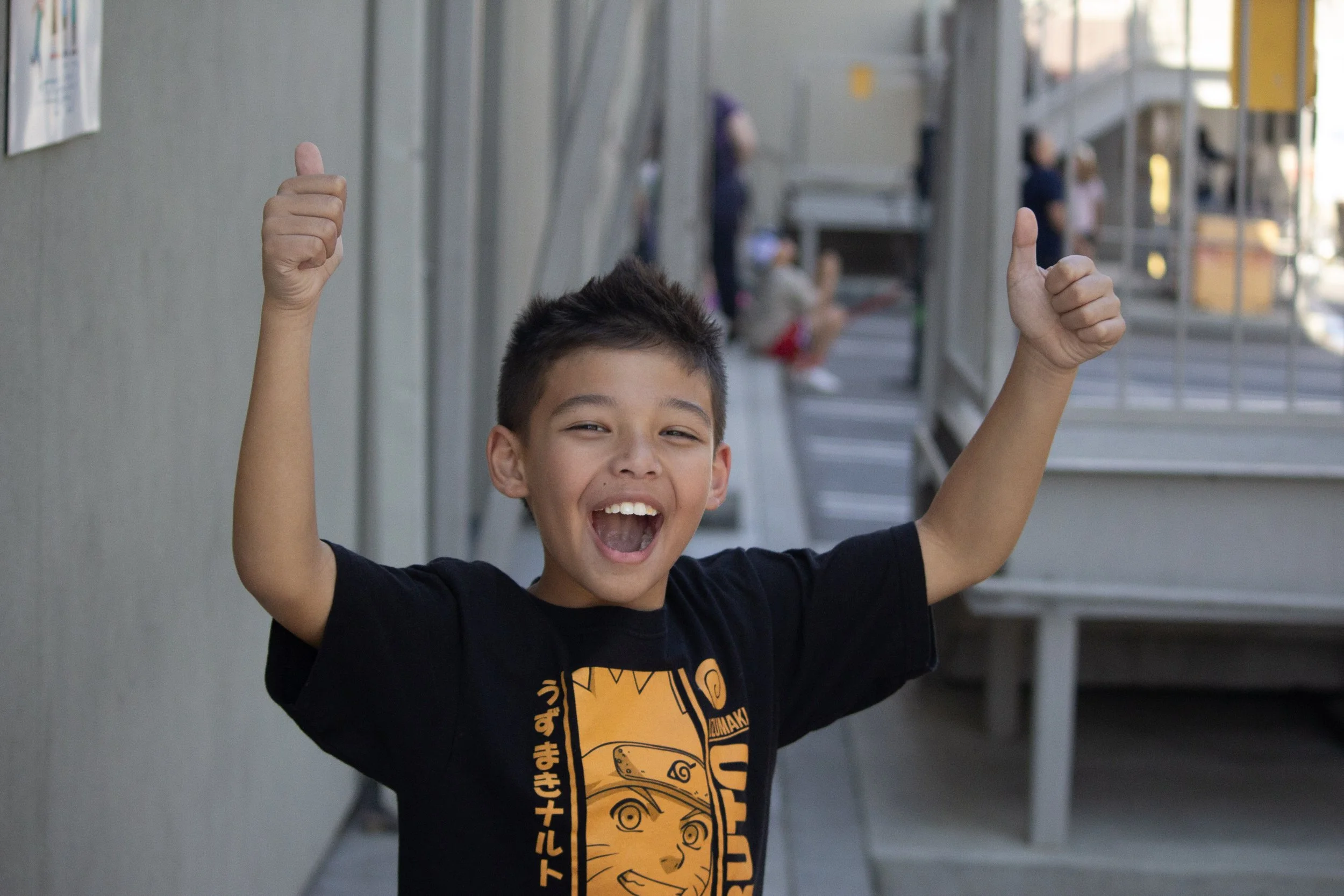 A young boy celebrating outdoors with his arms raised and thumbs up, smiling widely.