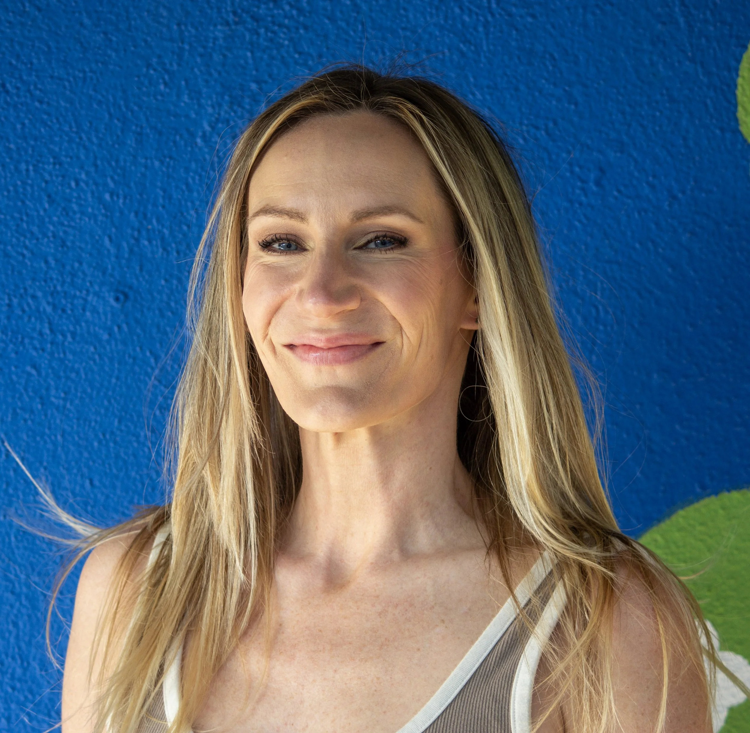 A woman with wavy blonde hair and blue eyes, smiling and wearing a sleeveless pink top, standing outdoors with blurred greenery in the background.
