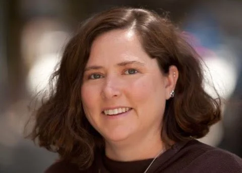 Close-up of a smiling woman with shoulder-length brown hair, wearing a dark top.