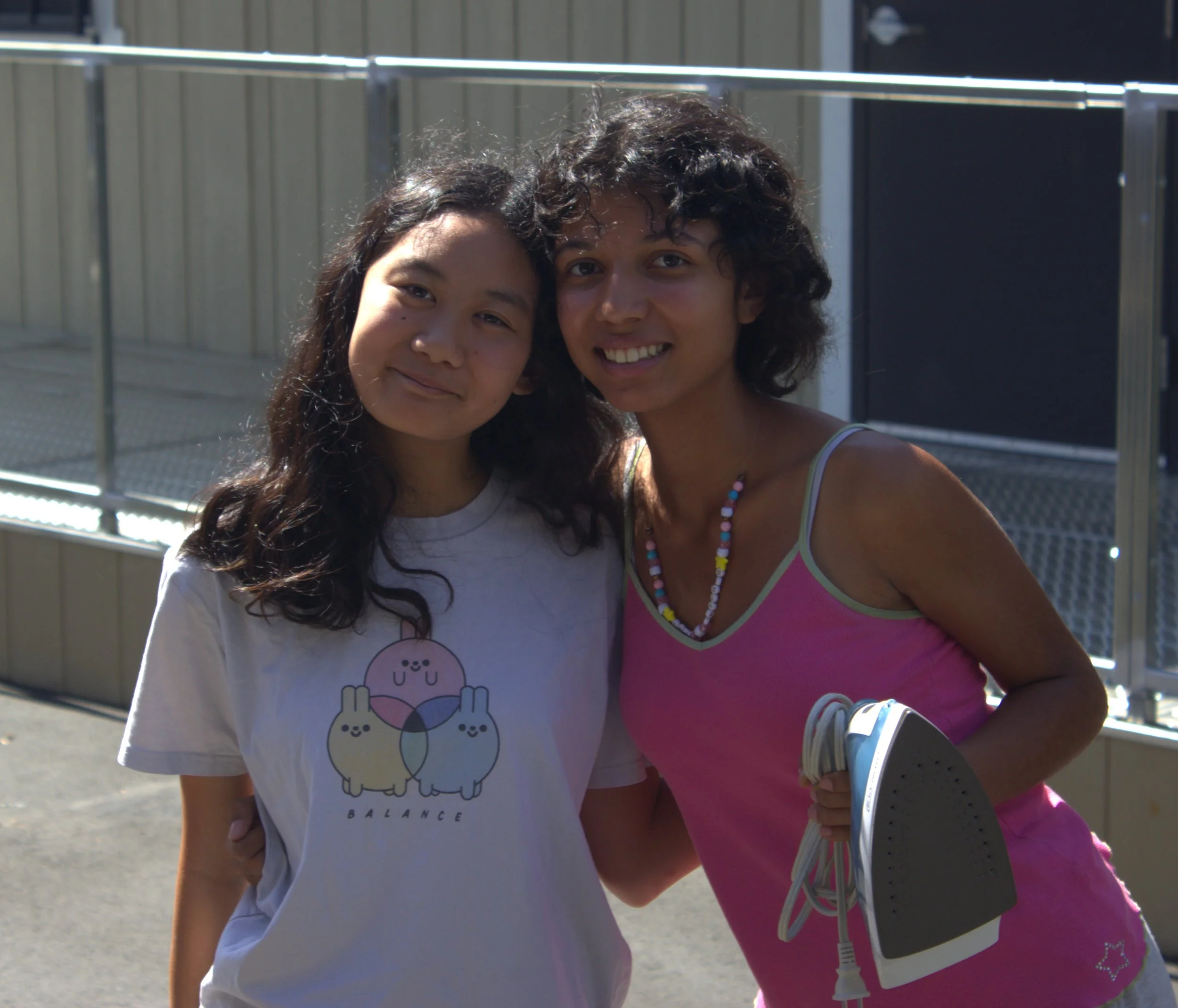 Two women standing close together, smiling outdoors on a sunny day, with a metal railing and building in the background.