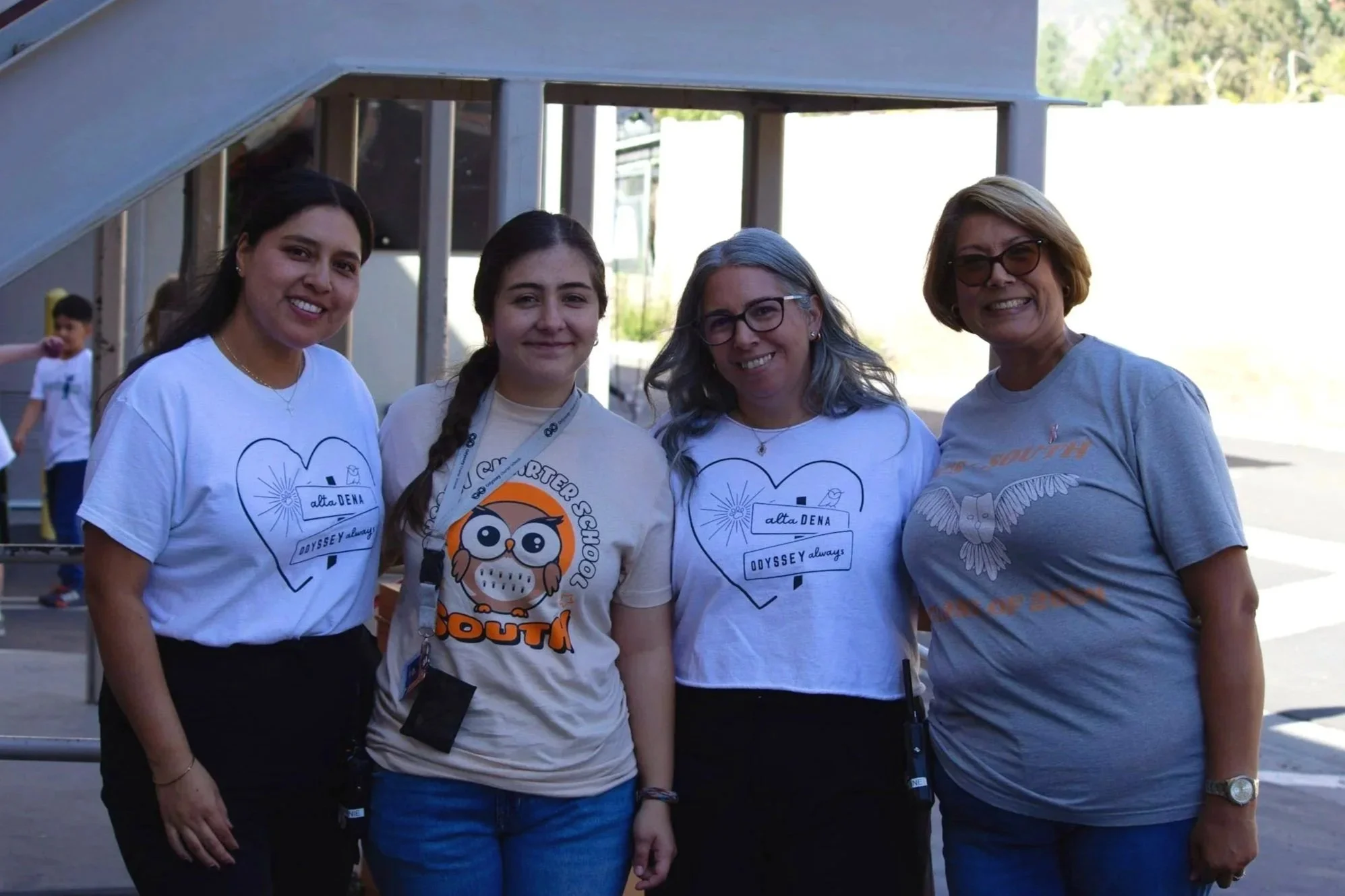 Four women standing outdoors, smiling at the camera, wearing shirts with event logos, with children playing in the background.
