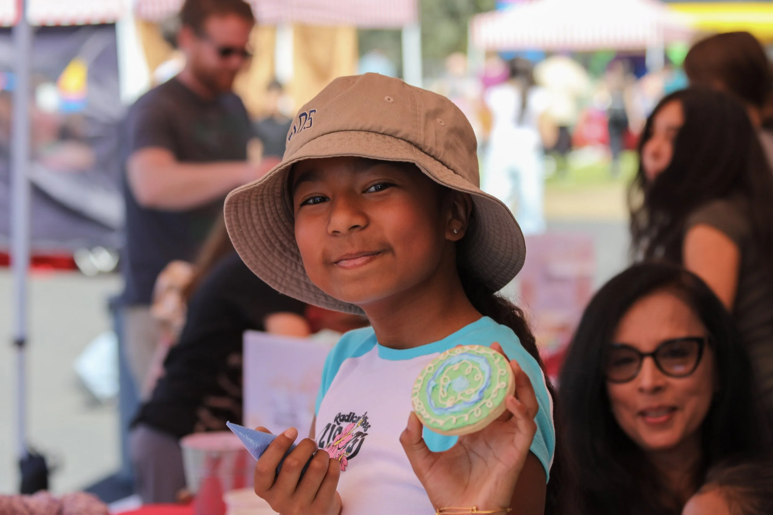 A young girl wearing a beige sun hat and a blue and white shirt holding a decorated cookie at an outdoor event.