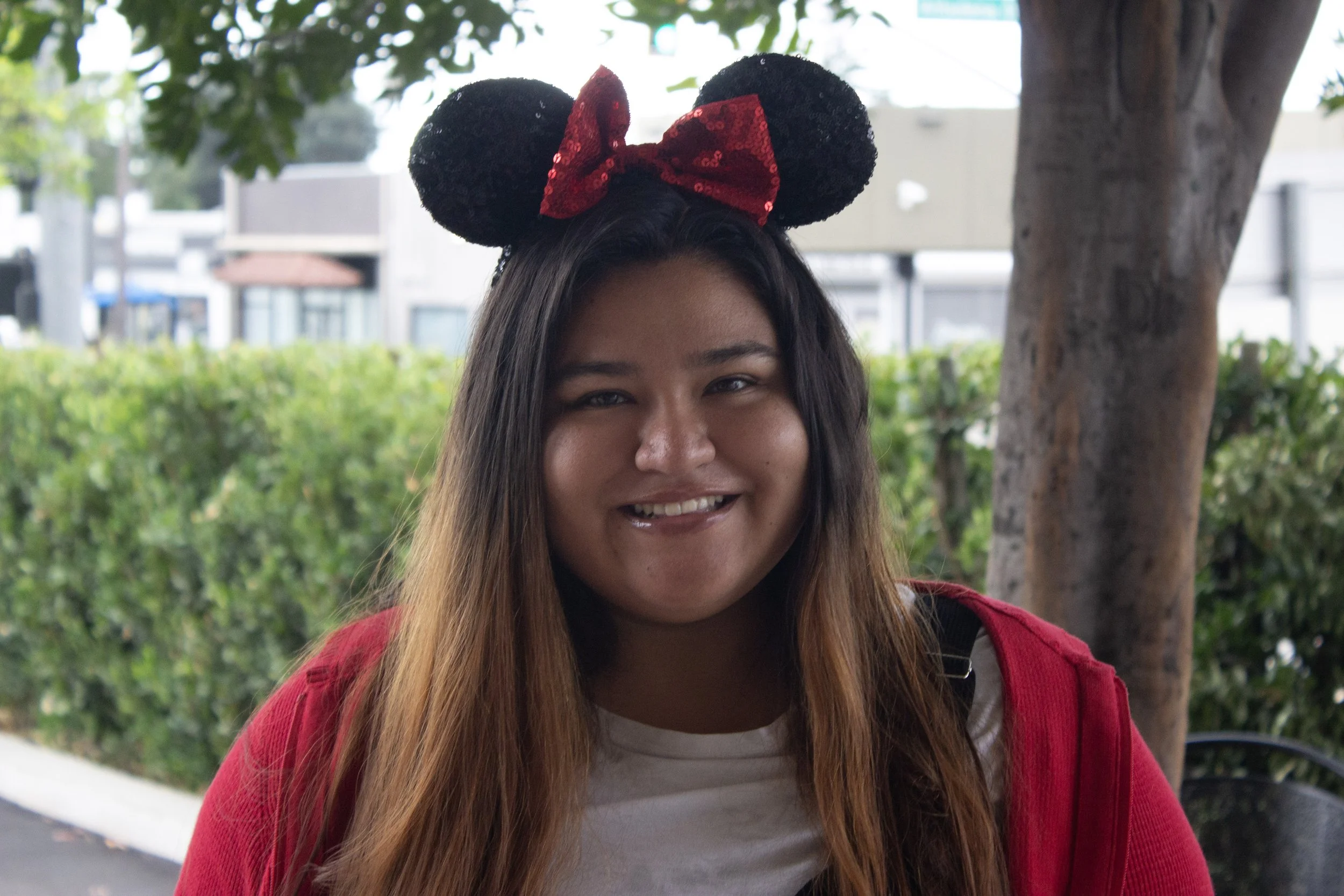 Woman wearing black and red sequin Minnie Mouse ears headband, smiling, outdoors near a tree with greenery and buildings in the background.