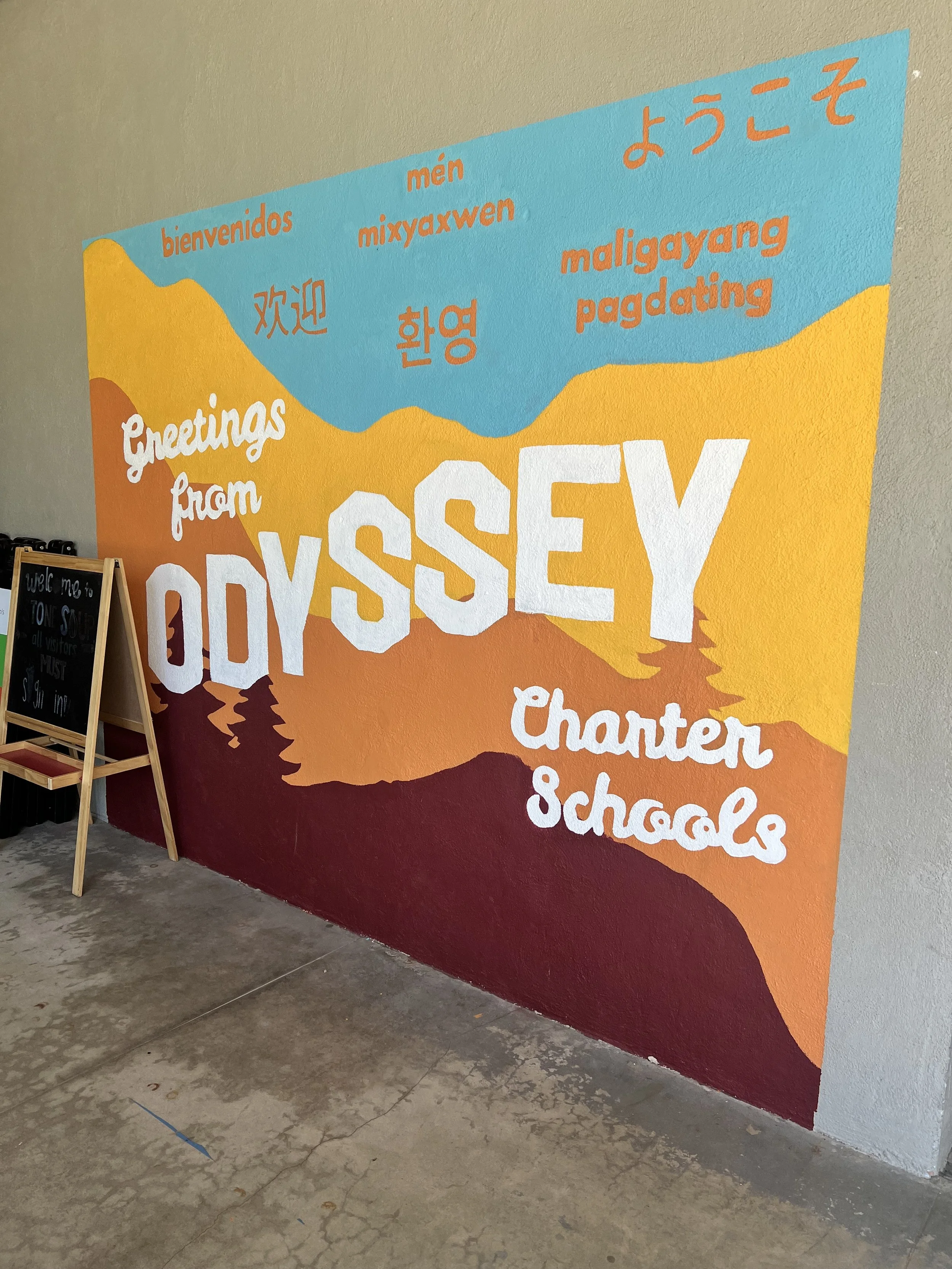 Colorful mural with text in multiple languages greeting from Odyssey Charter Schools, featuring mountains and trees in a landscape scene.