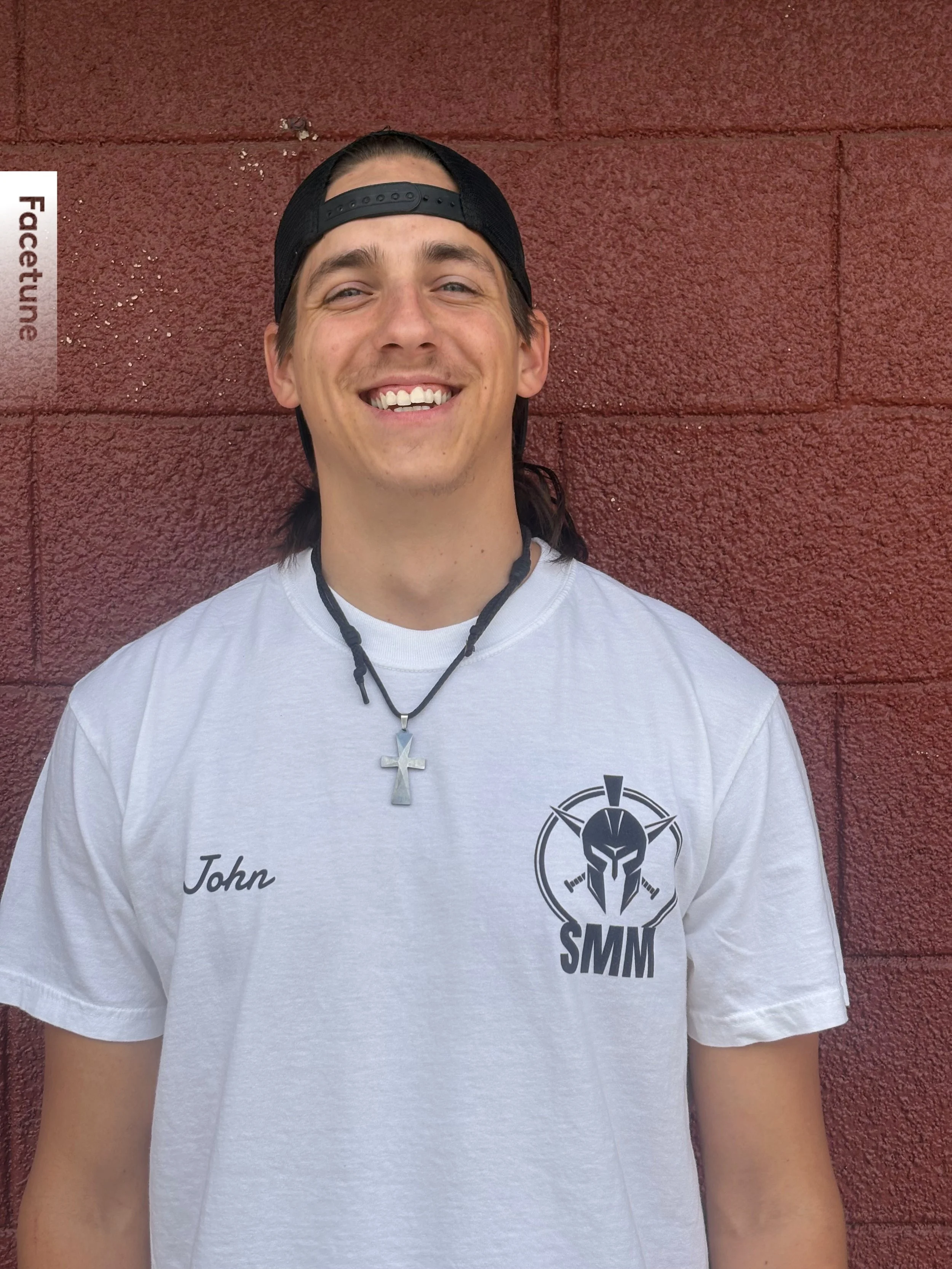 Smiling young man with long hair, wearing a black cap backward, a necklace with a silver cross, a white T-shirt with a logo and the name "John" on it, standing in front of a red textured wall.