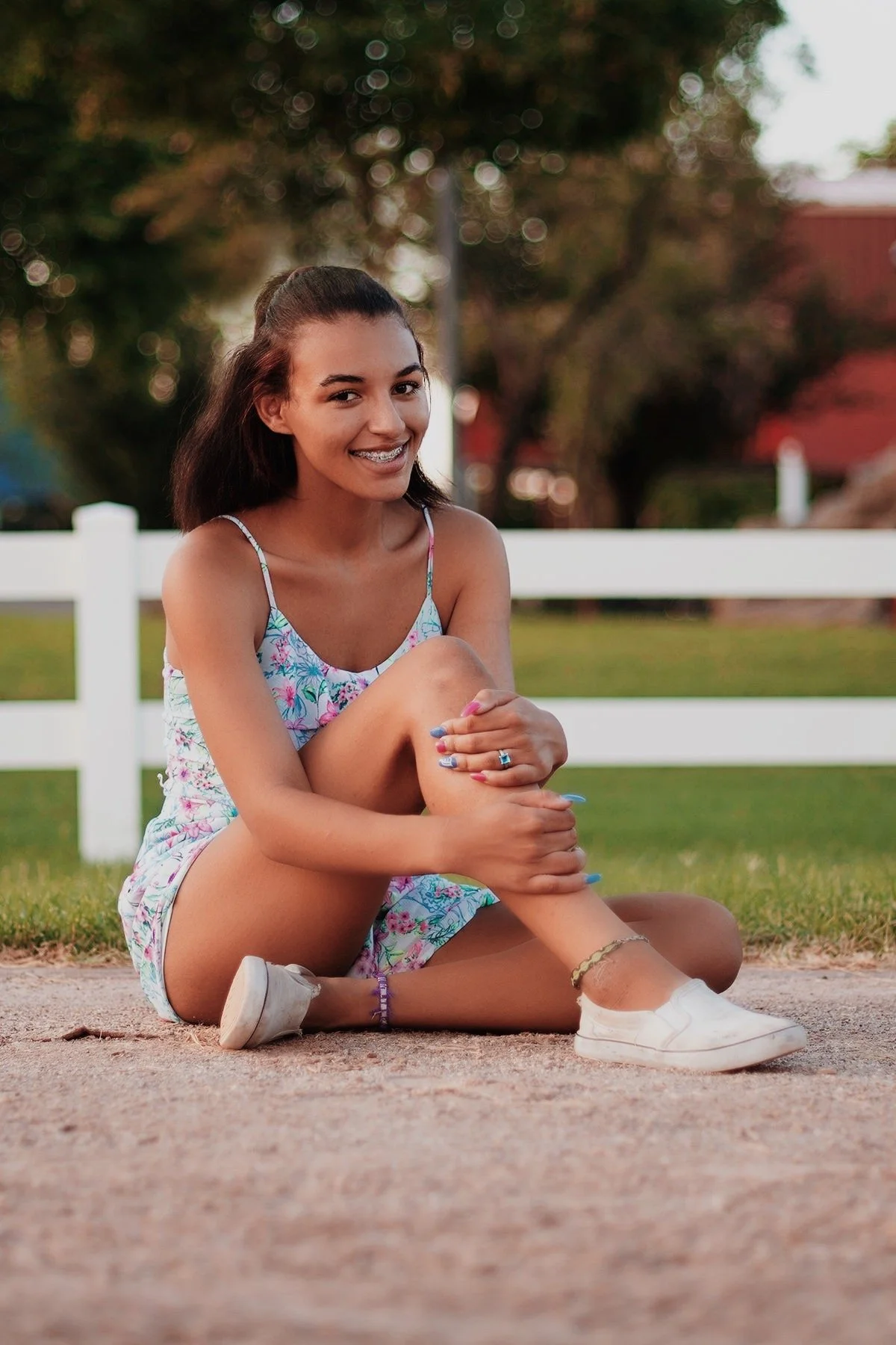 A young woman sitting on the ground outdoors, smiling, wearing a colorful floral dress, white sneakers, and jewelry. She is holding her leg with one hand and has braces on her teeth. There are trees and a white fence in the background.