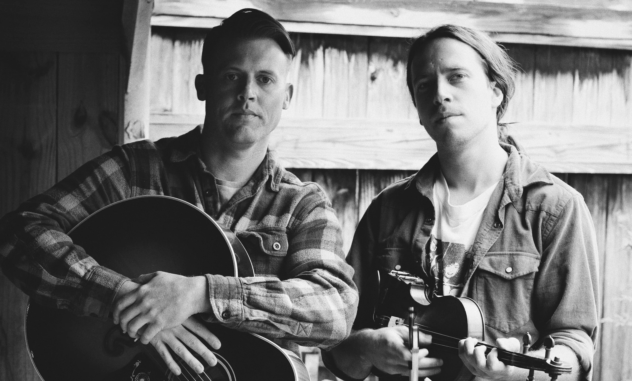 Two young men in casual clothing holding acoustic guitars, standing near a wooden wall, looking at the camera in a black and white photograph.