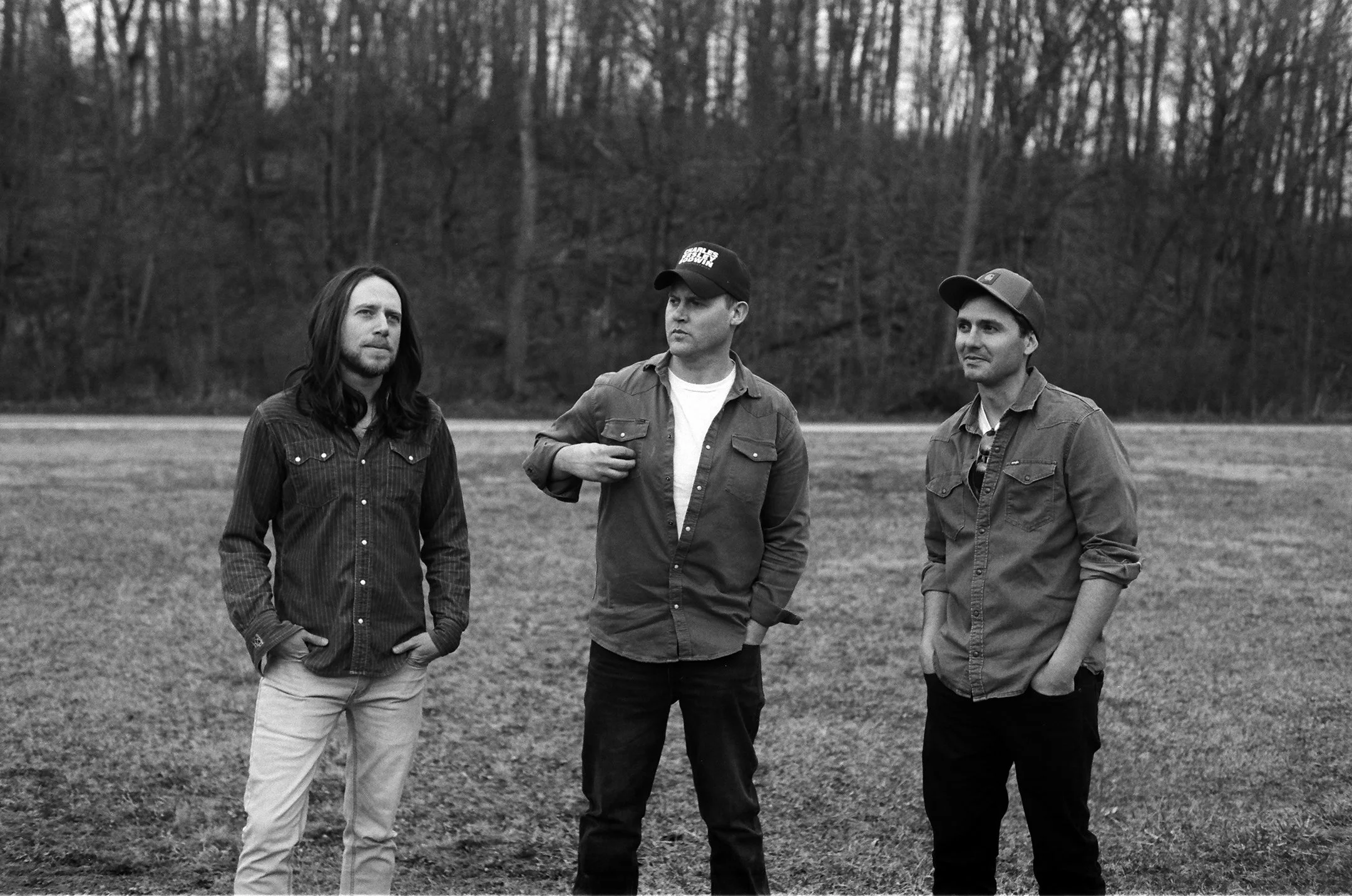 Three men standing outdoors on a grassy field with trees in the background, wearing casual jackets and hats, in black and white.