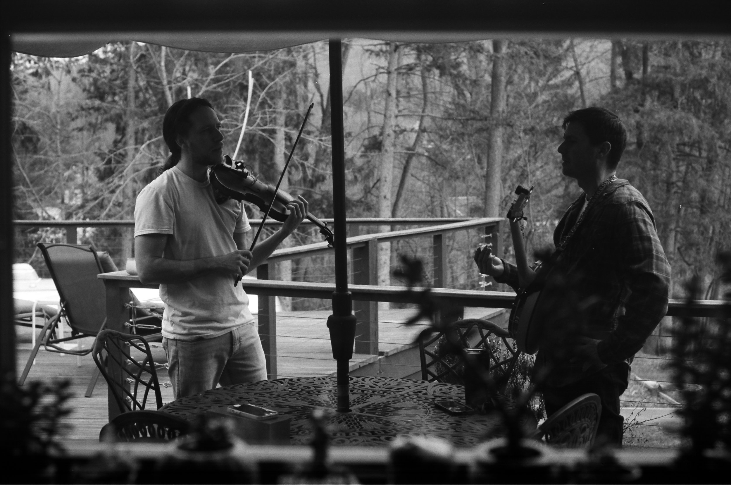 Two men playing musical instruments on a wooden deck outdoors, viewed through a window with blinds. One man is playing a violin and the other is holding a guitar.