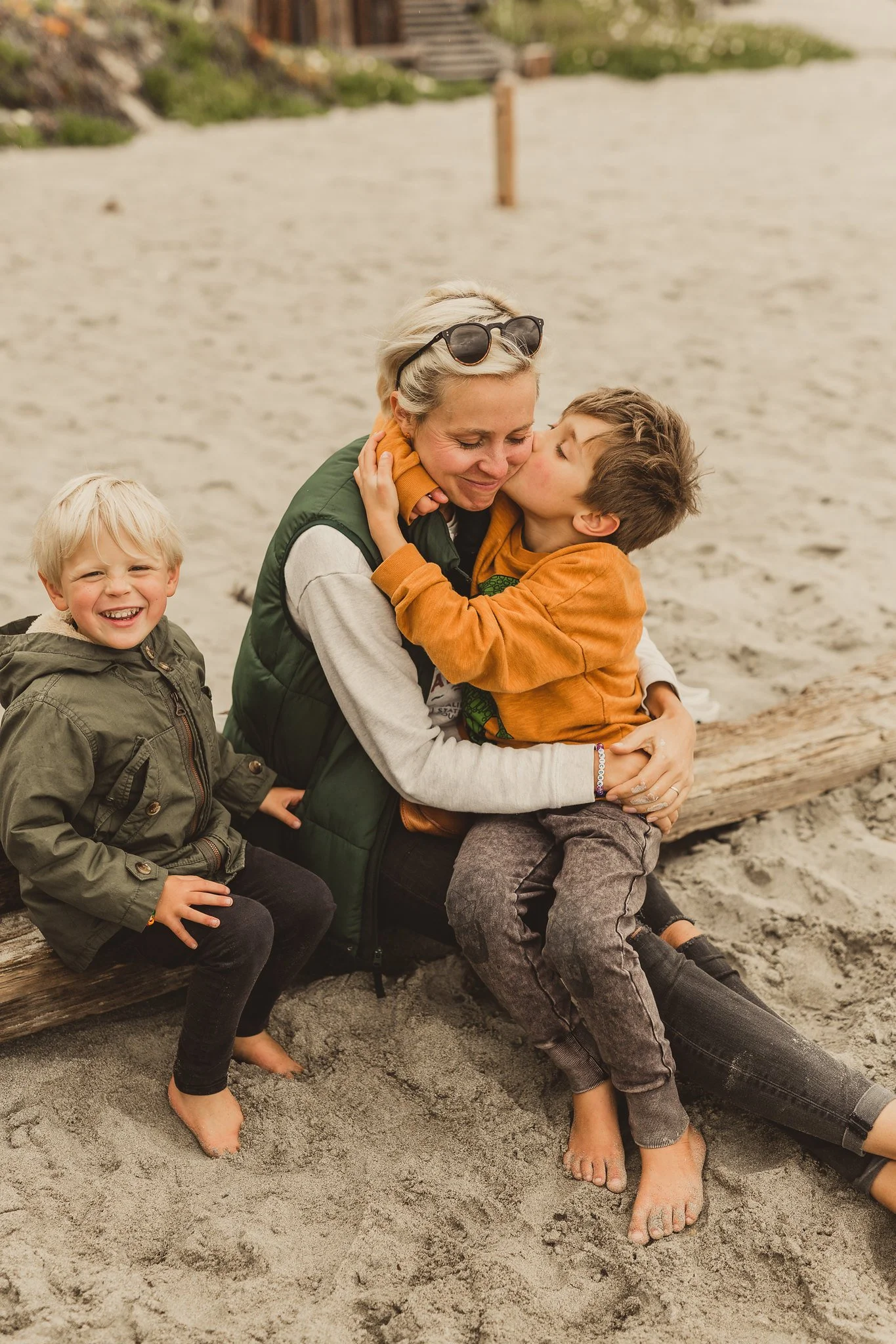 A woman with short blonde hair hugging a young boy who is kissing her cheek at the beach. Another smiling boy sits beside them, all barefoot on the sand with a driftwood log behind them.