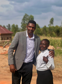 A man and a woman standing outdoors. The man has his arm around the woman, and they are smiling. The background includes a house, trees, and a cloudy sky.