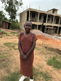 Young girl standing outdoors in front of an unfinished building and trees, wearing a brown dress.