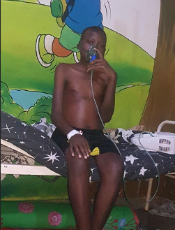 A shirtless boy sitting on a hospital bed holding a nebulizer mask to his face in a room with colorful cartoon wall decor.