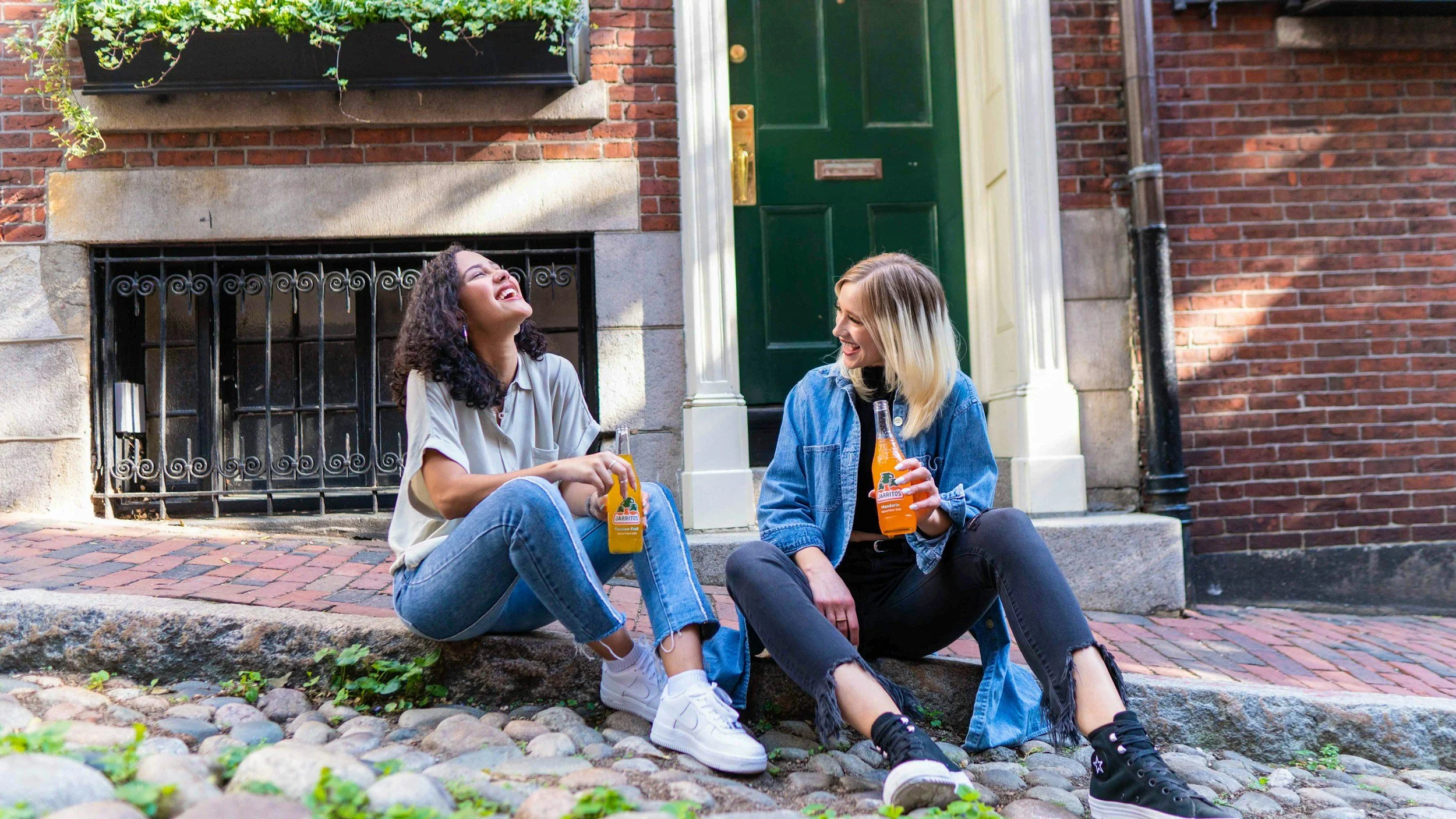 Two young women sitting on a cobblestone sidewalk, laughing and holding bottles of orange soda, outside a house with a green door and brick exterior.