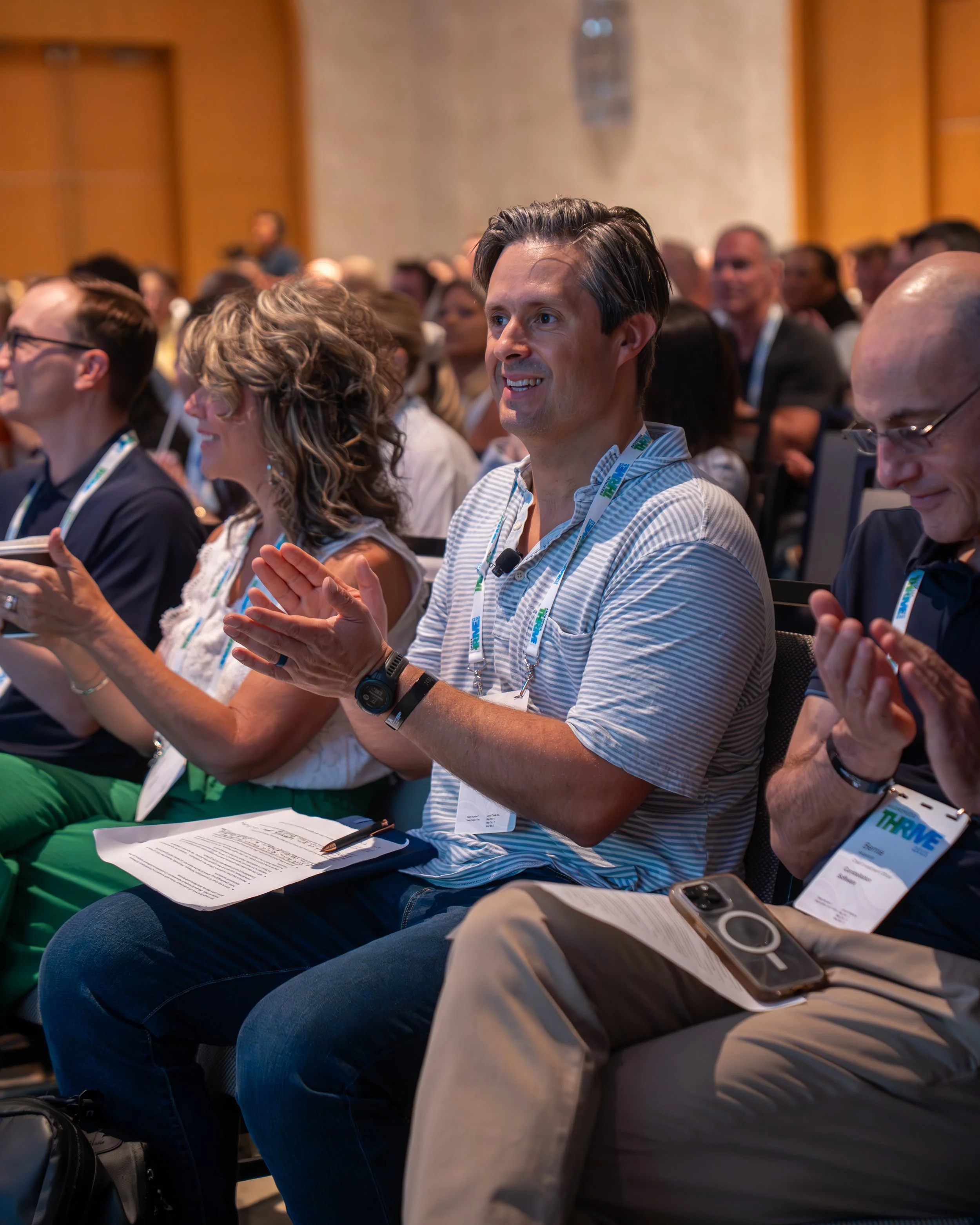 Attendees at a conference sitting in rows, clapping and smiling, wearing conference badges around their necks.