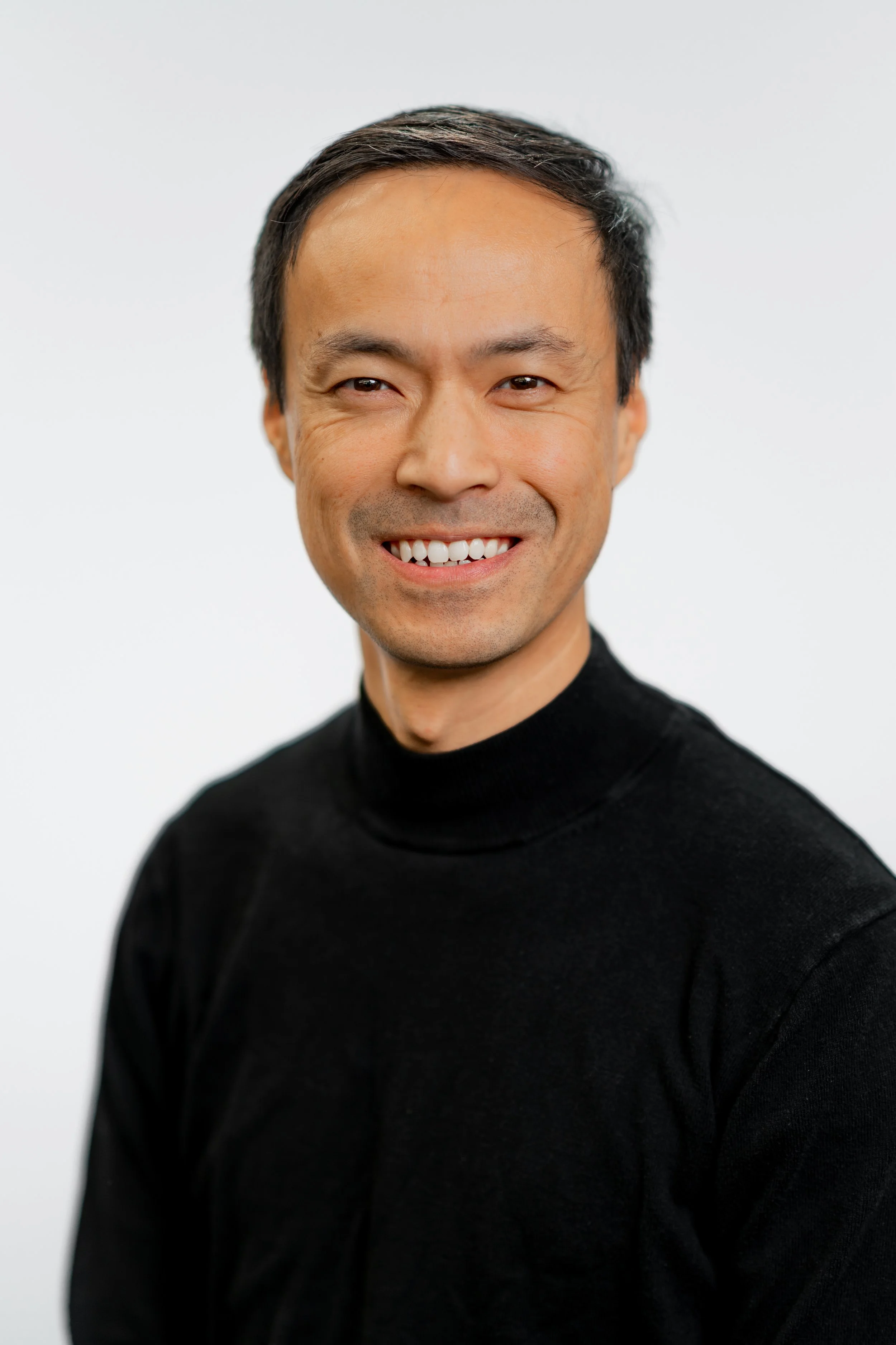 A smiling man with short dark hair wearing a black turtleneck sweater, posing against a plain white background.
