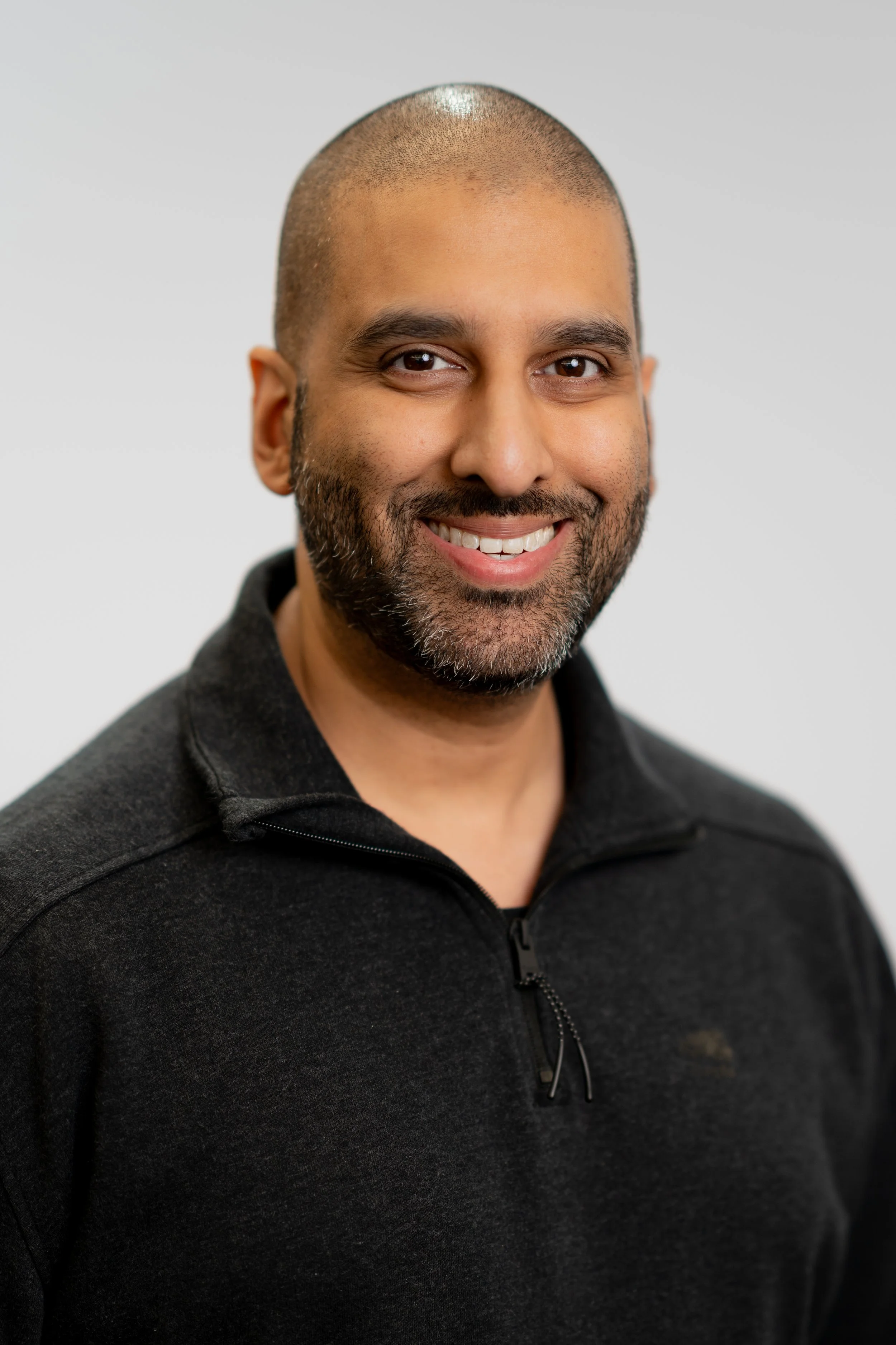 Headshot of a smiling man with a beard, wearing a black zip-up fleece jacket, against a plain light background.