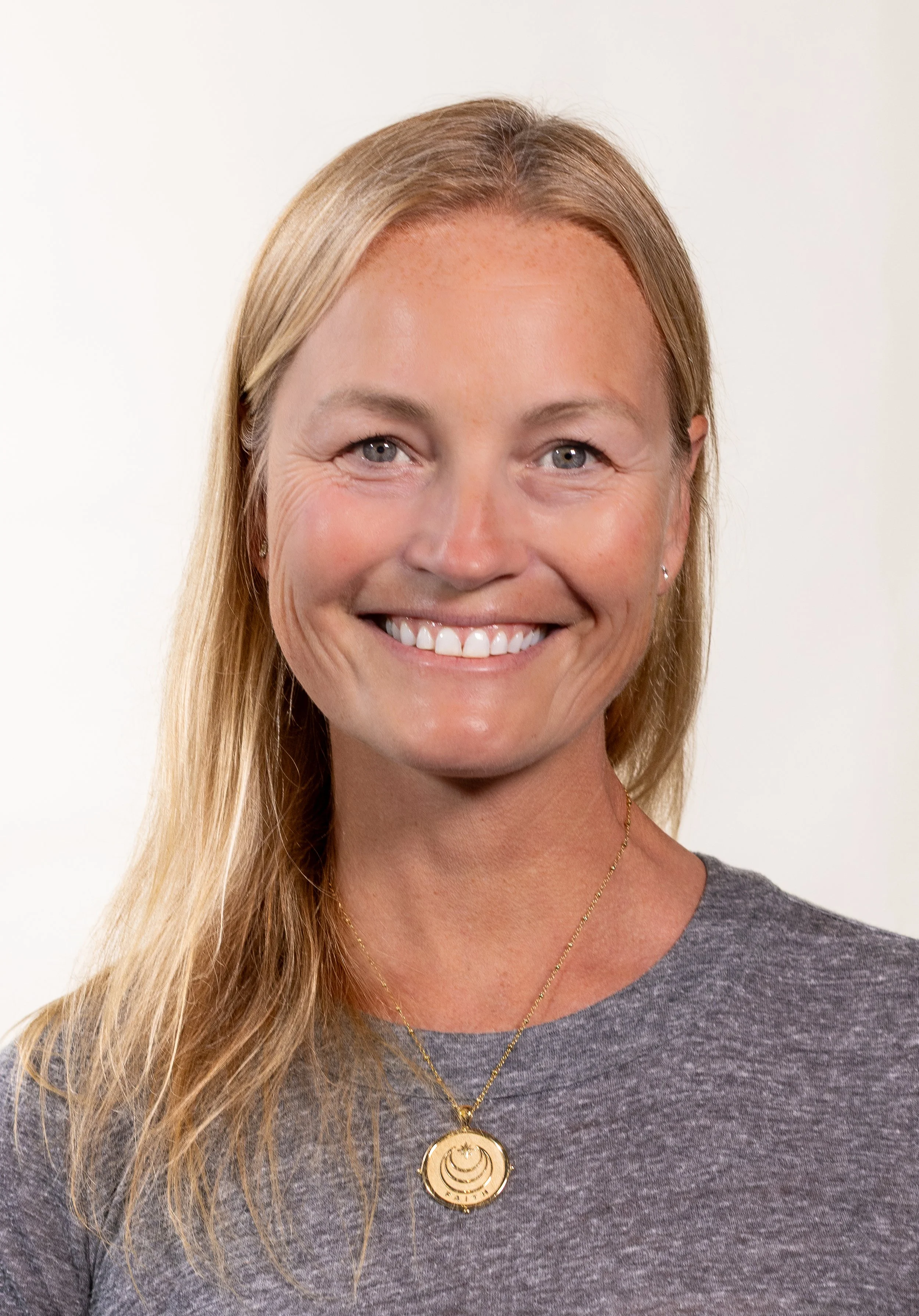 A woman with blonde hair, blue eyes, and a smile, wearing a gray shirt and a gold necklace, standing against a plain background.