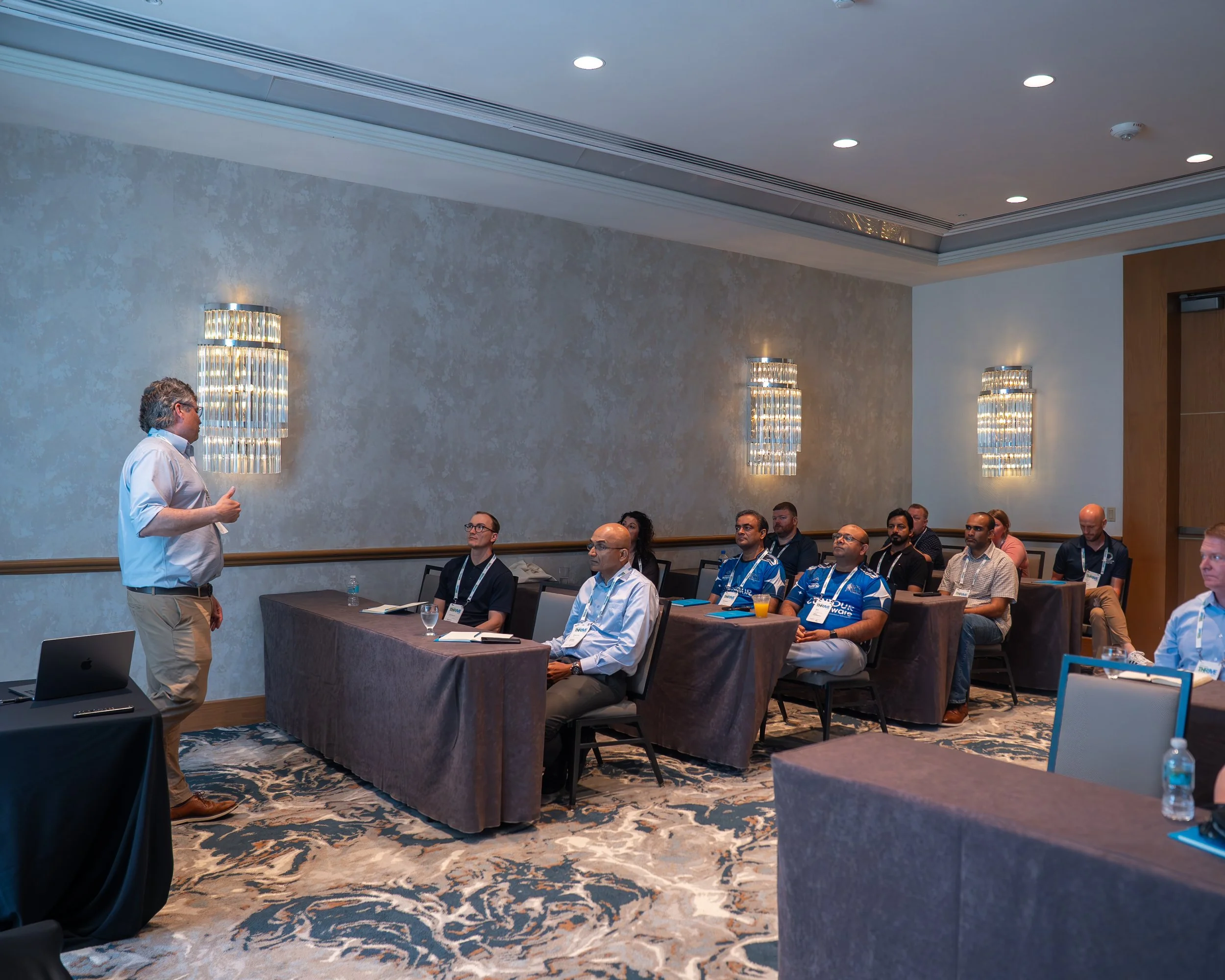 A man giving a presentation to an audience seated at tables in a conference room with beige walls and modern light fixtures.