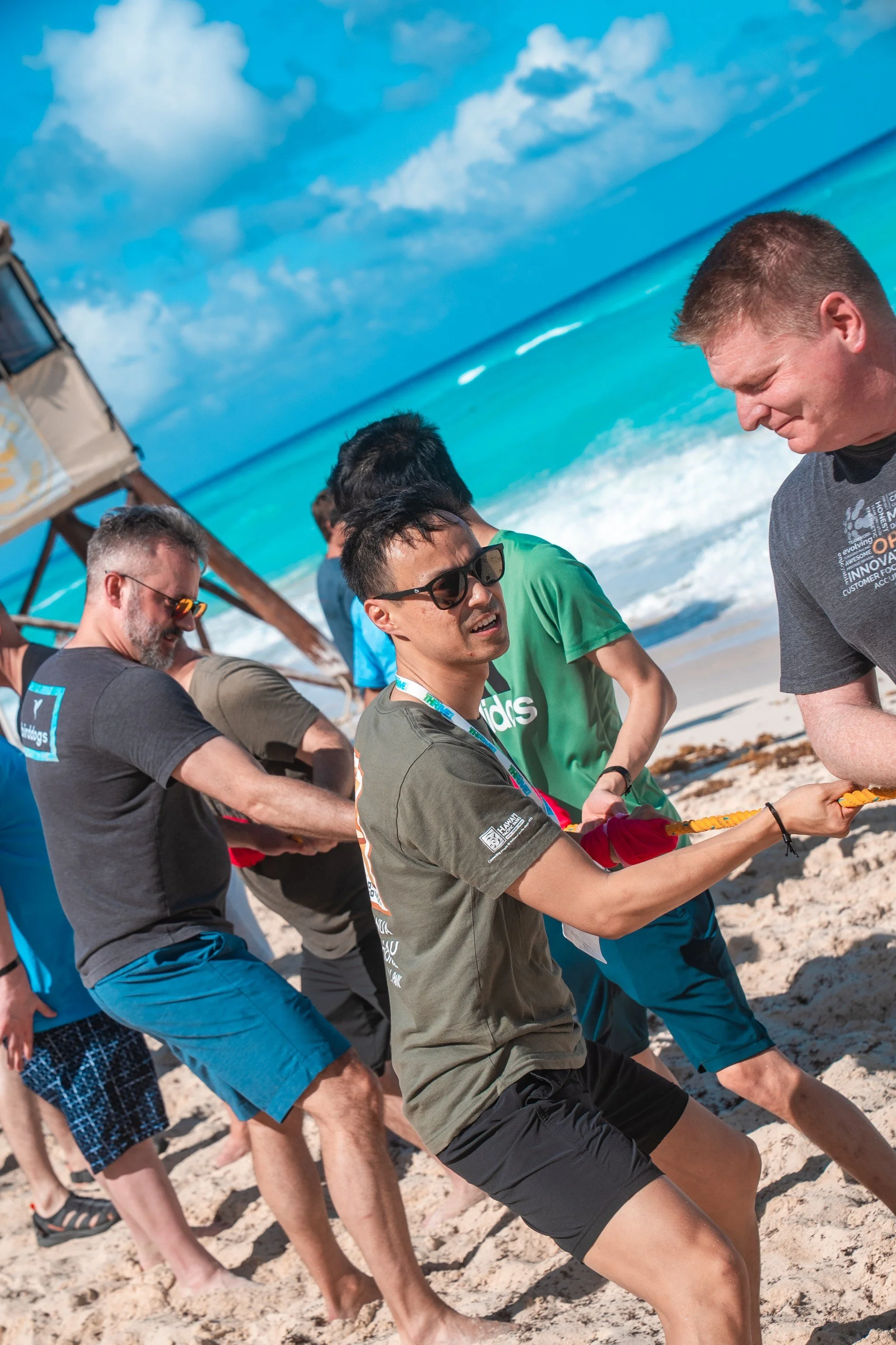 Group of people participating in a tug-of-war game on a sandy beach with ocean and blue sky in the background.