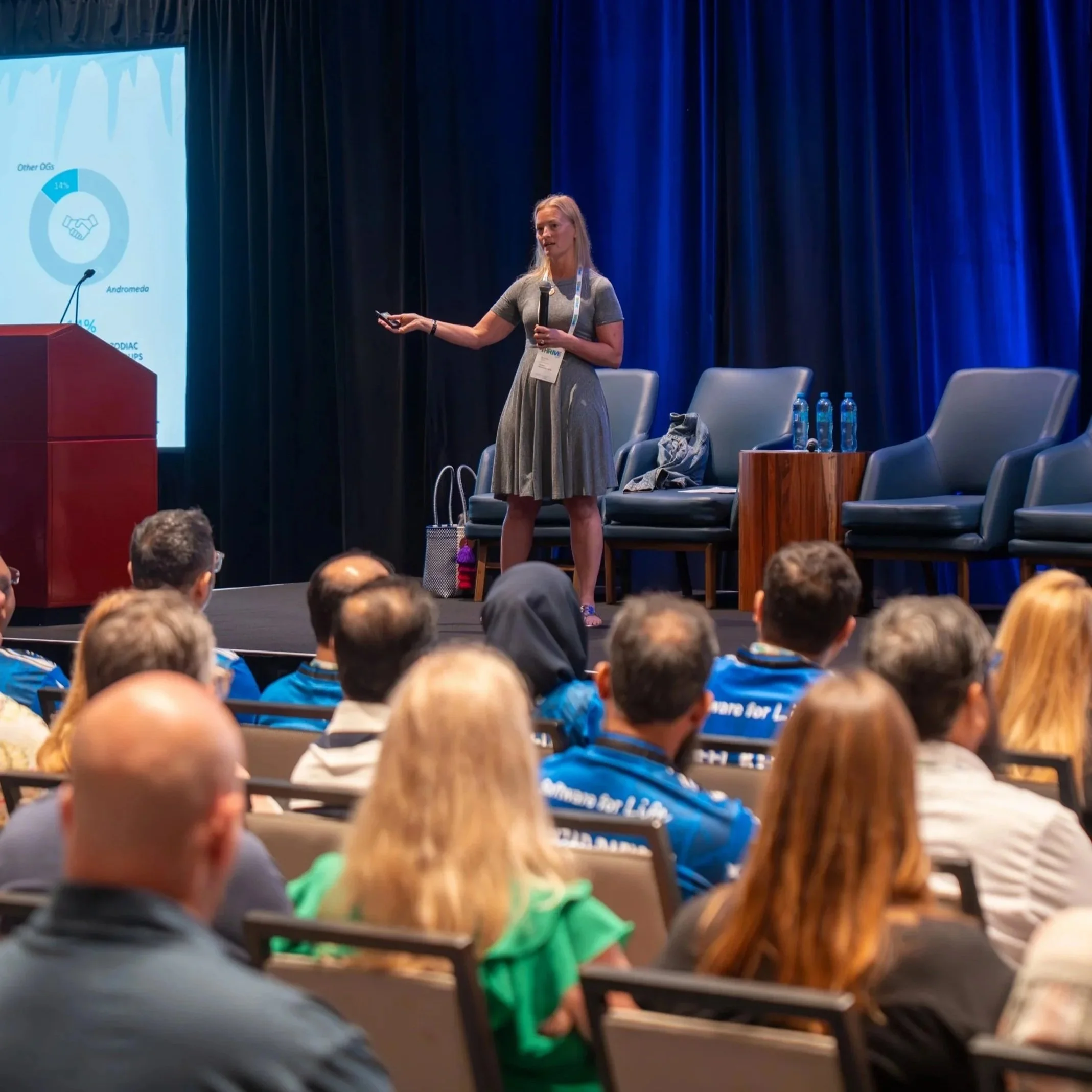A woman in a gray dress giving a presentation on stage at a conference, with a large screen displaying a chart behind her and an audience seated facing her.