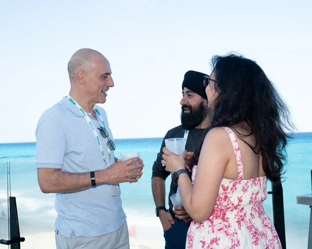 Three people are smiling and talking at the beach, holding drinks. Two men and one woman, with ocean waves in the background.