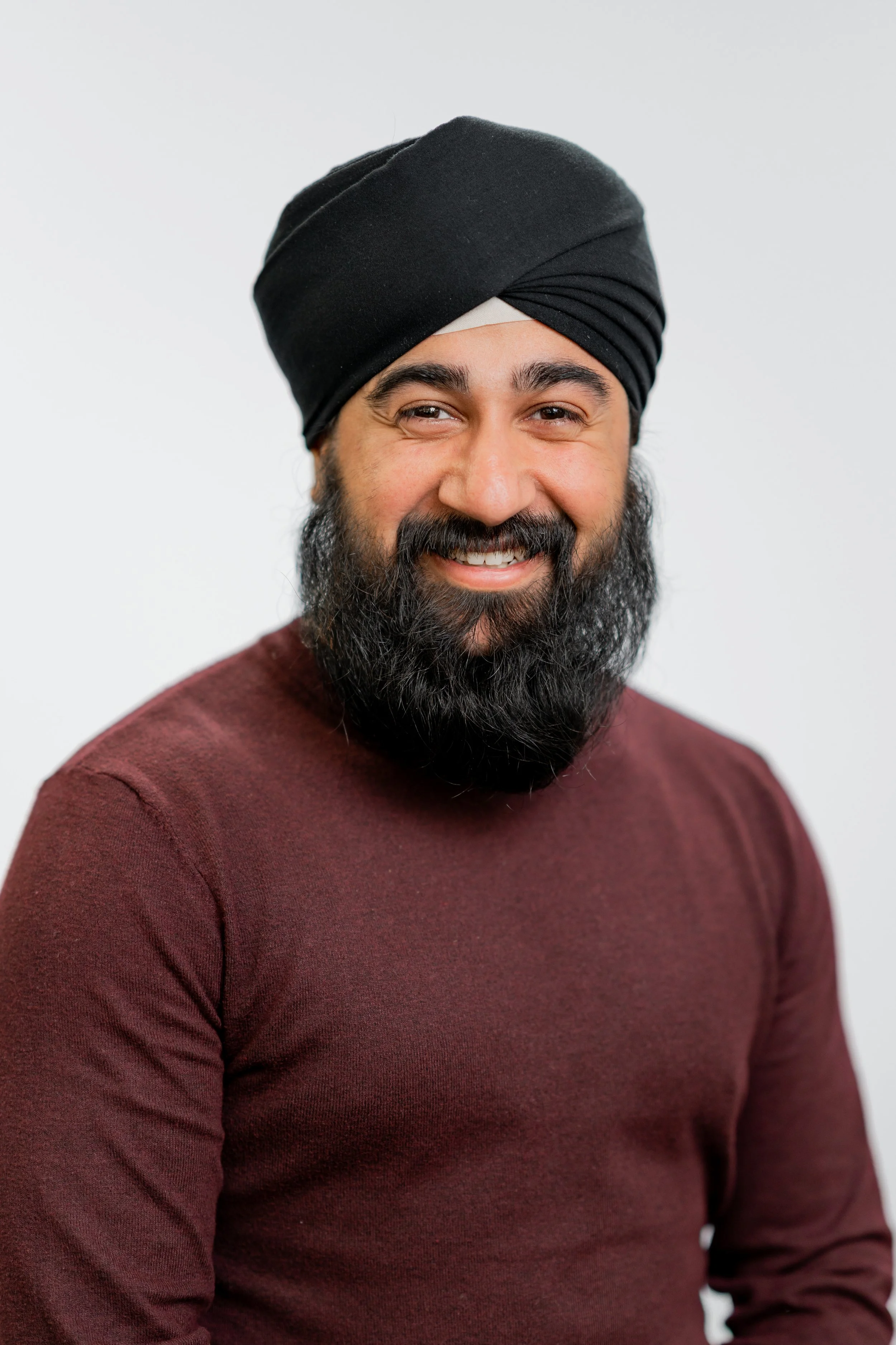 Portrait of a man with a dark beard and mustache, wearing a black turban and a maroon shirt, smiling against a plain white background.
