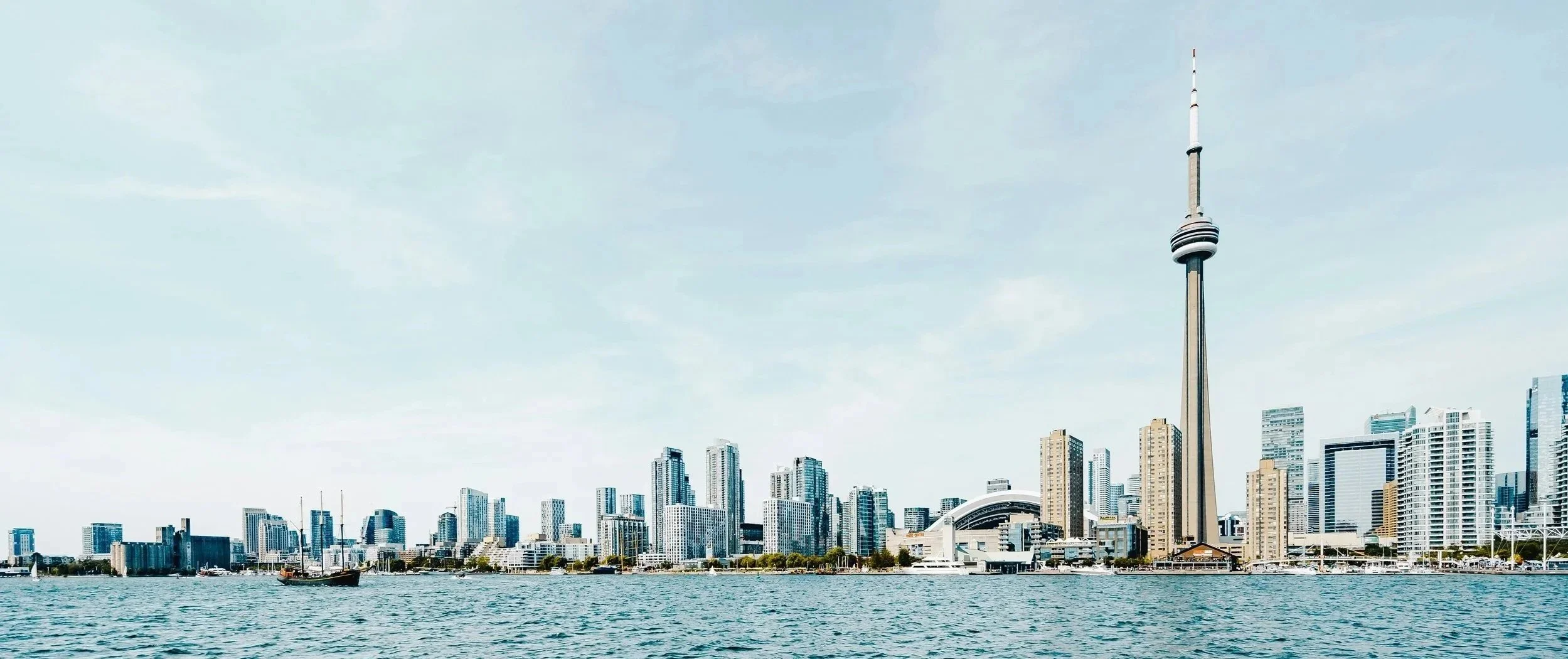A city skyline featuring the CN Tower and various high-rise buildings viewed across a body of water with a small boat.