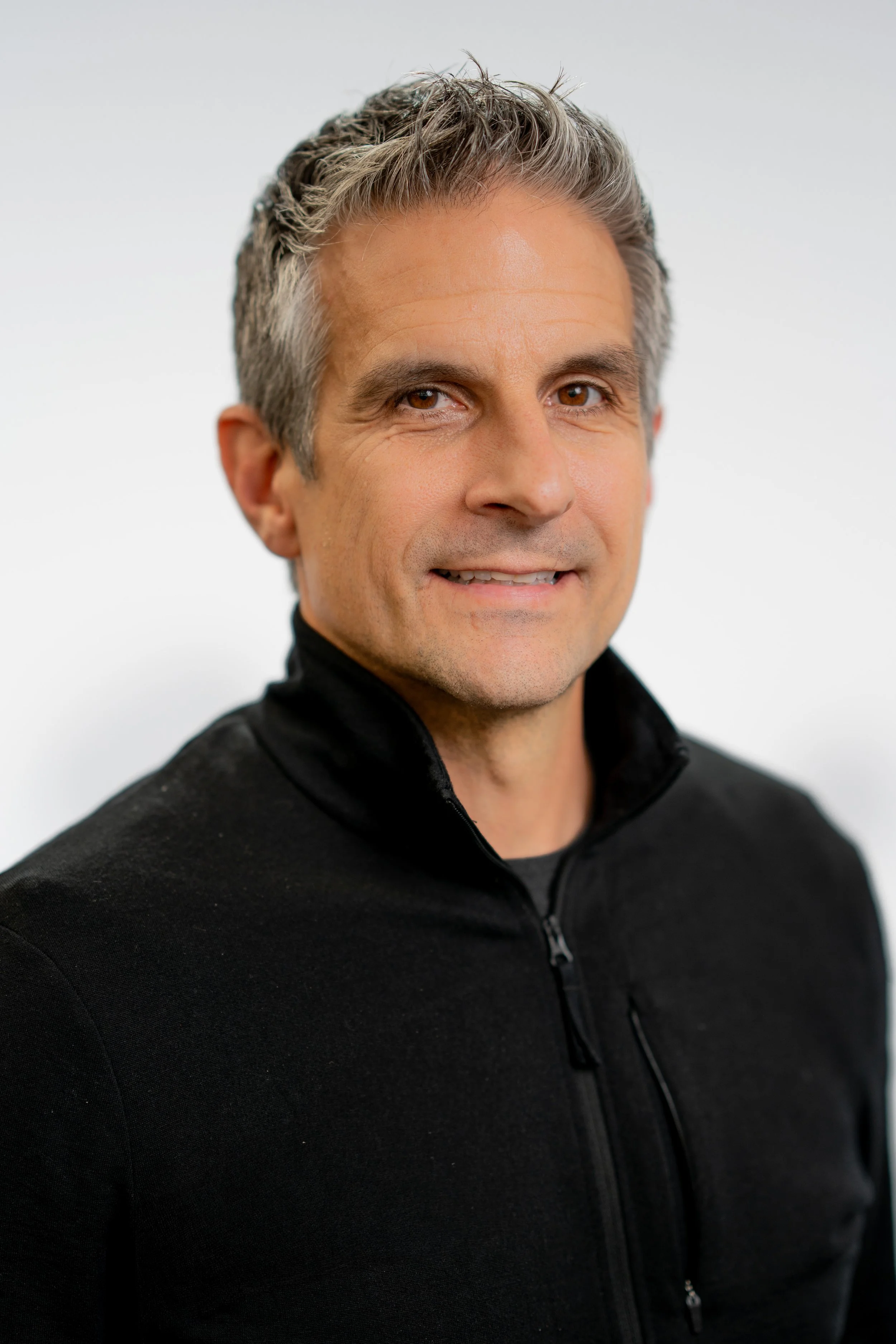 Portrait of a middle-aged man with short, styled gray hair, wearing a black zip-up jacket, smiling slightly against a plain white background.