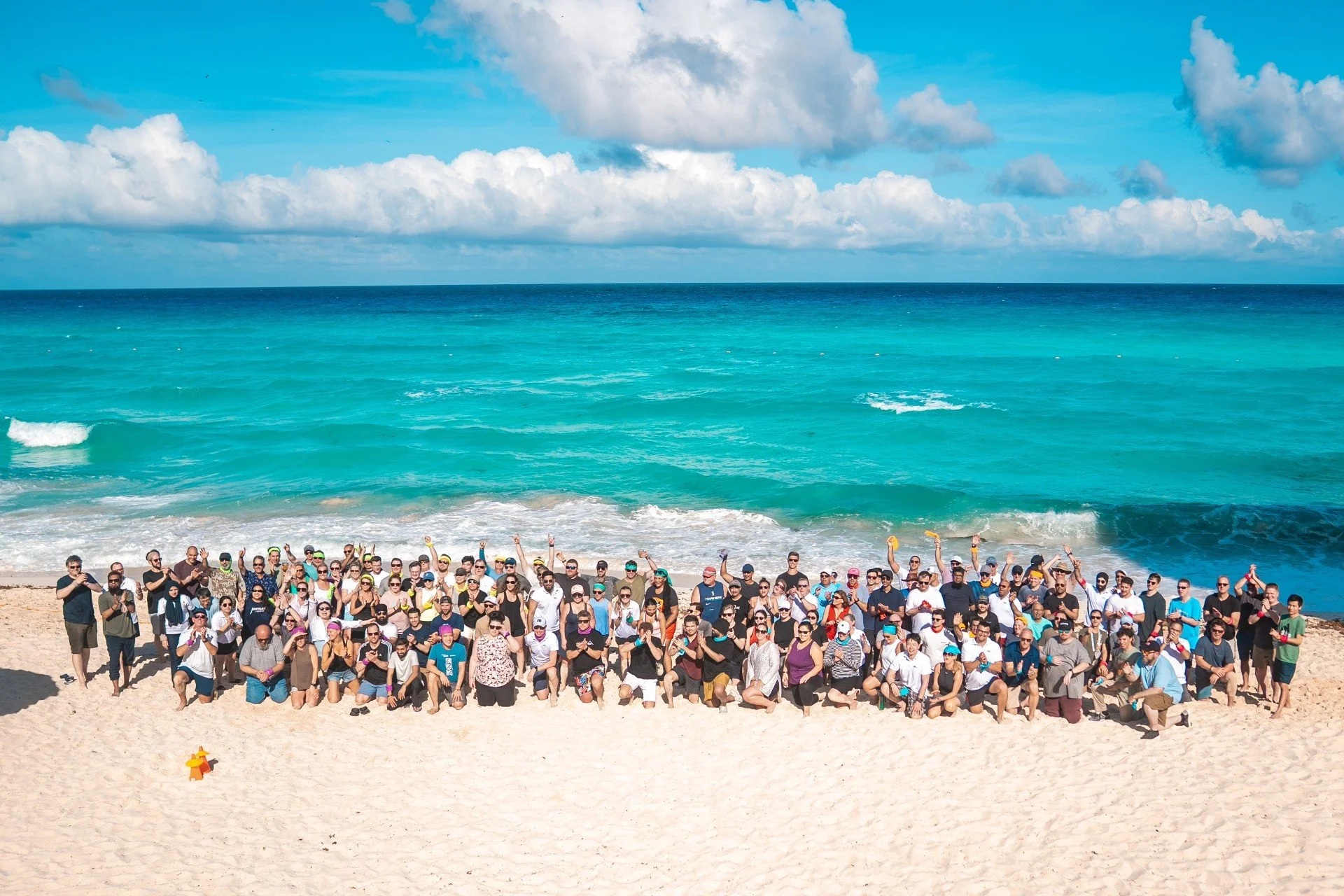 Large group of people gathered on a sandy beach near the turquoise ocean, with blue sky and white clouds overhead.