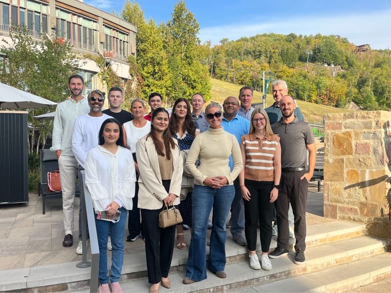Group of 14 people standing outdoors on a sunny day with trees and a hillside in the background.