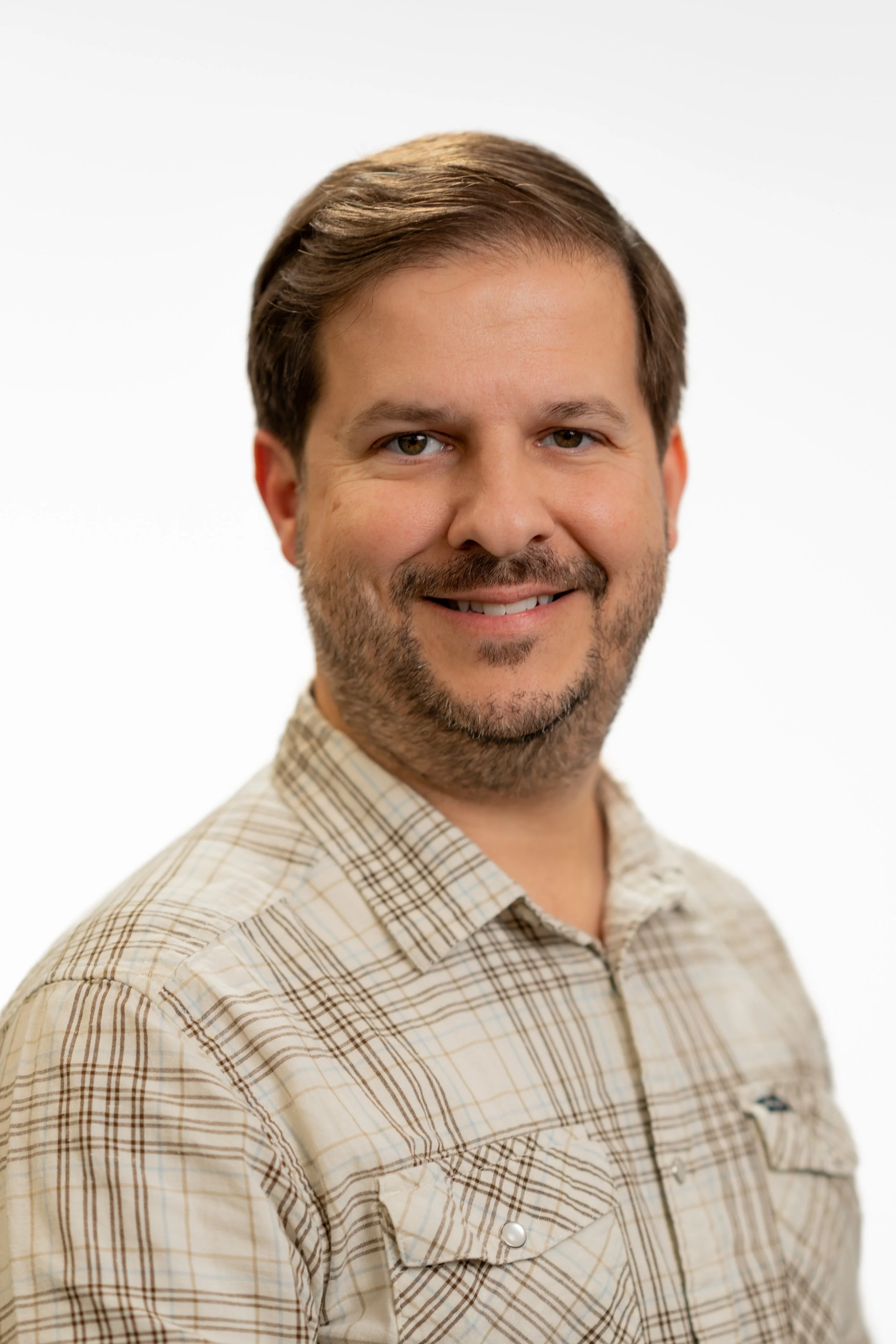 Portrait of a man with brown hair, beard, and mustache, smiling, wearing a plaid beige shirt, against a plain white background.