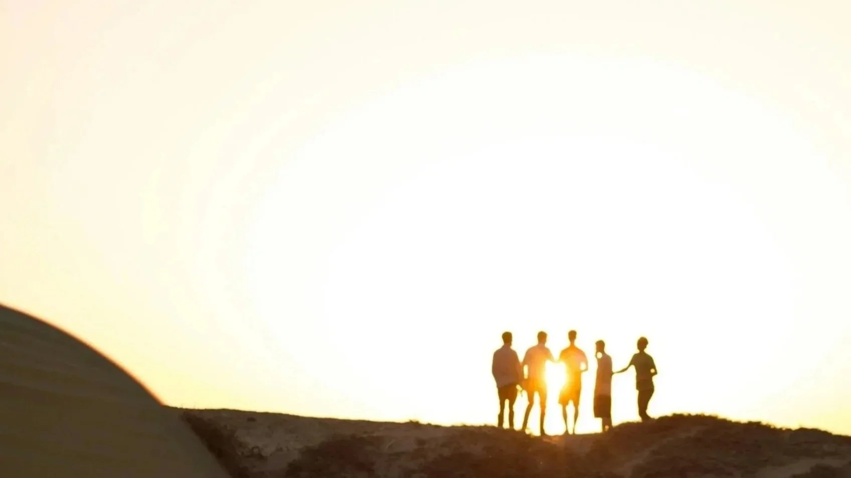 Group of six people standing together on a hilltop during sunset, silhouetted against the bright sky, some holding hands.