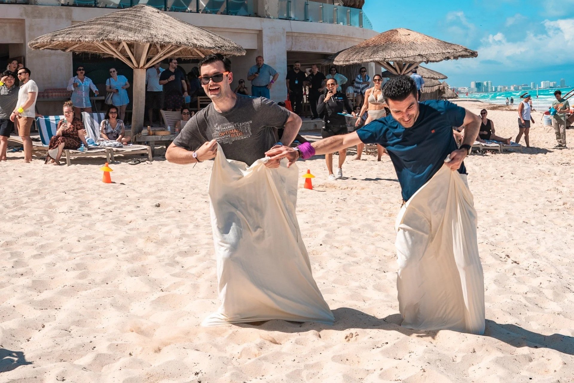 Two men participating in a beach sack race, smiling and holding onto the top of their sacks, with a crowd of spectators and beach umbrellas in the background.