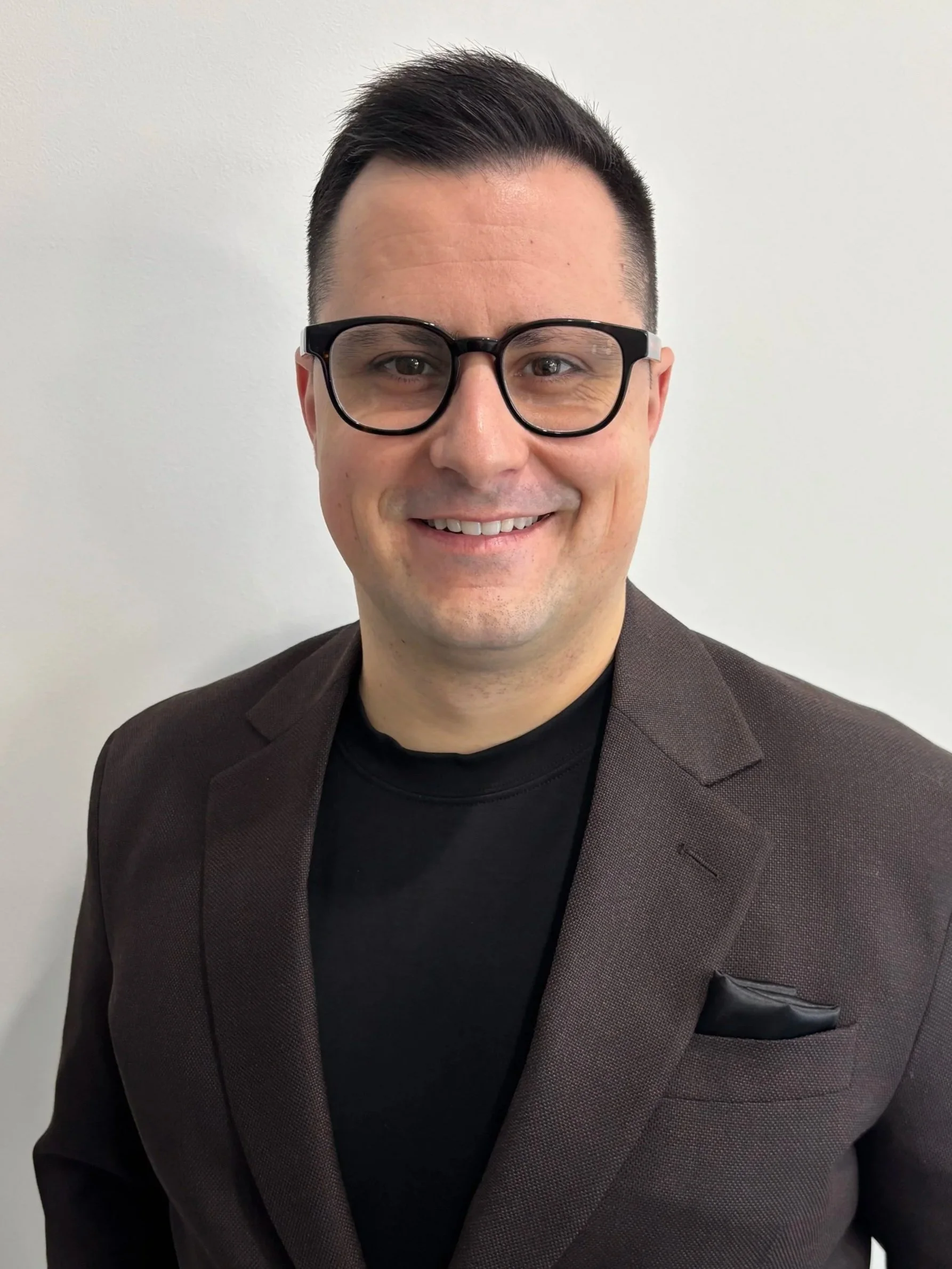 A smiling man with black hair wearing glasses, a black shirt, and a dark brown blazer with a pocket square stands against a plain white background.