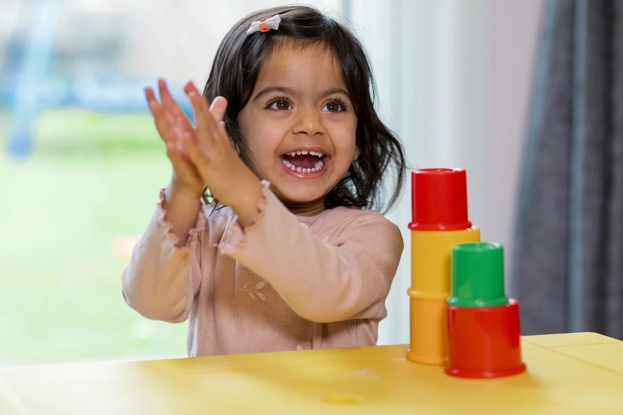 A young girl with dark hair in a pink shirt claps her hands as she plays with colorful stacking cups during communication therapy for kids in Las Vegas, NV