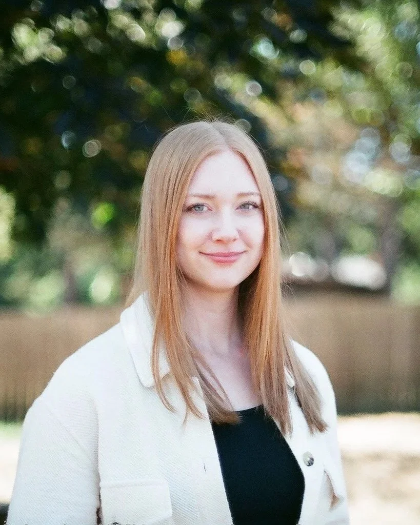 A young woman with straight, shoulder-length red hair, standing outdoors in a sunny setting with blurred trees and a fence in the background, smiling softly at the camera.