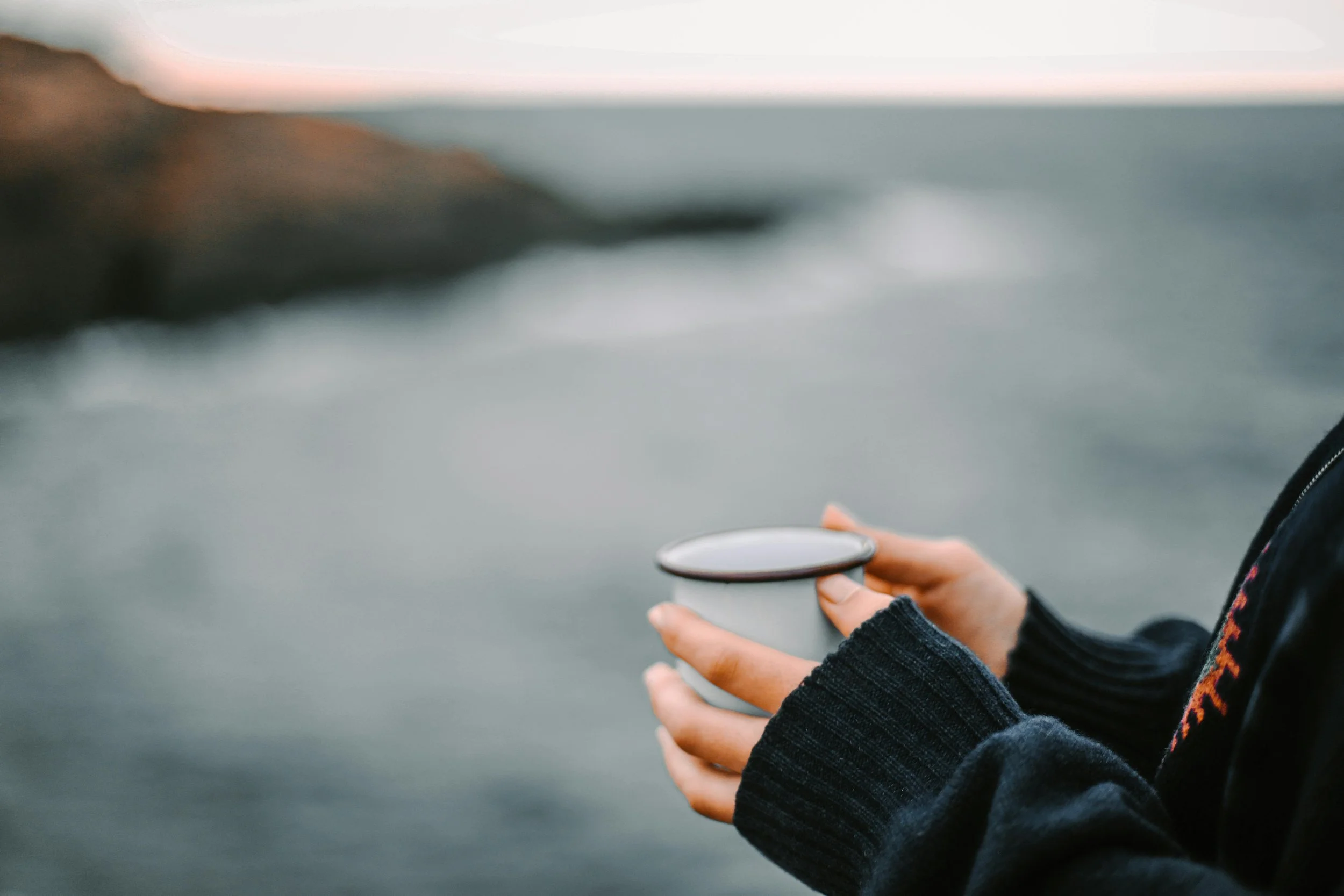 A person holding a coffee mug by the ocean, wearing a black sweater with a colorful pattern on the sleeve.