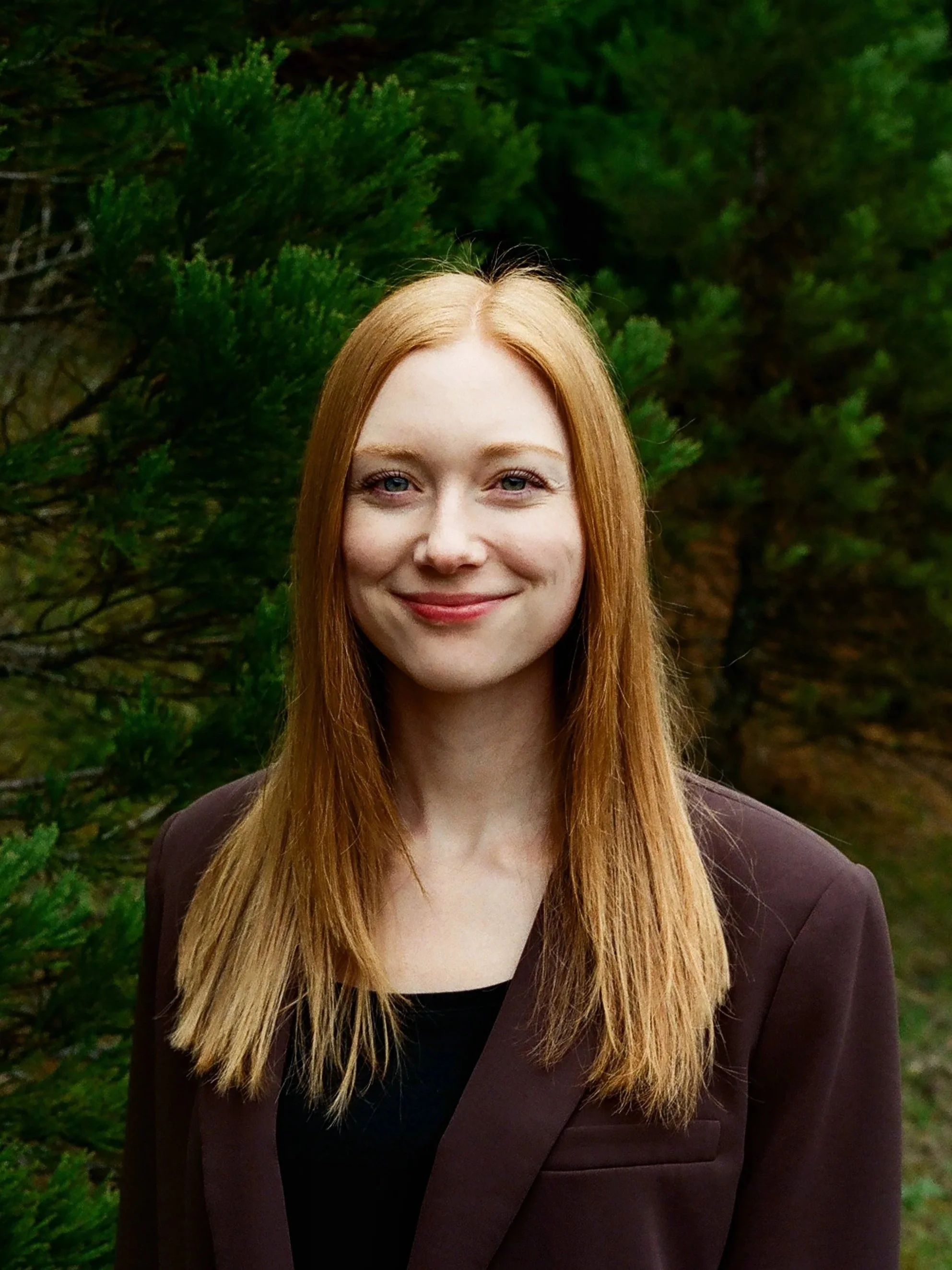 Portrait of a young woman with long red hair, smiling, wearing a dark blazer, standing outdoors with green foliage in the background.