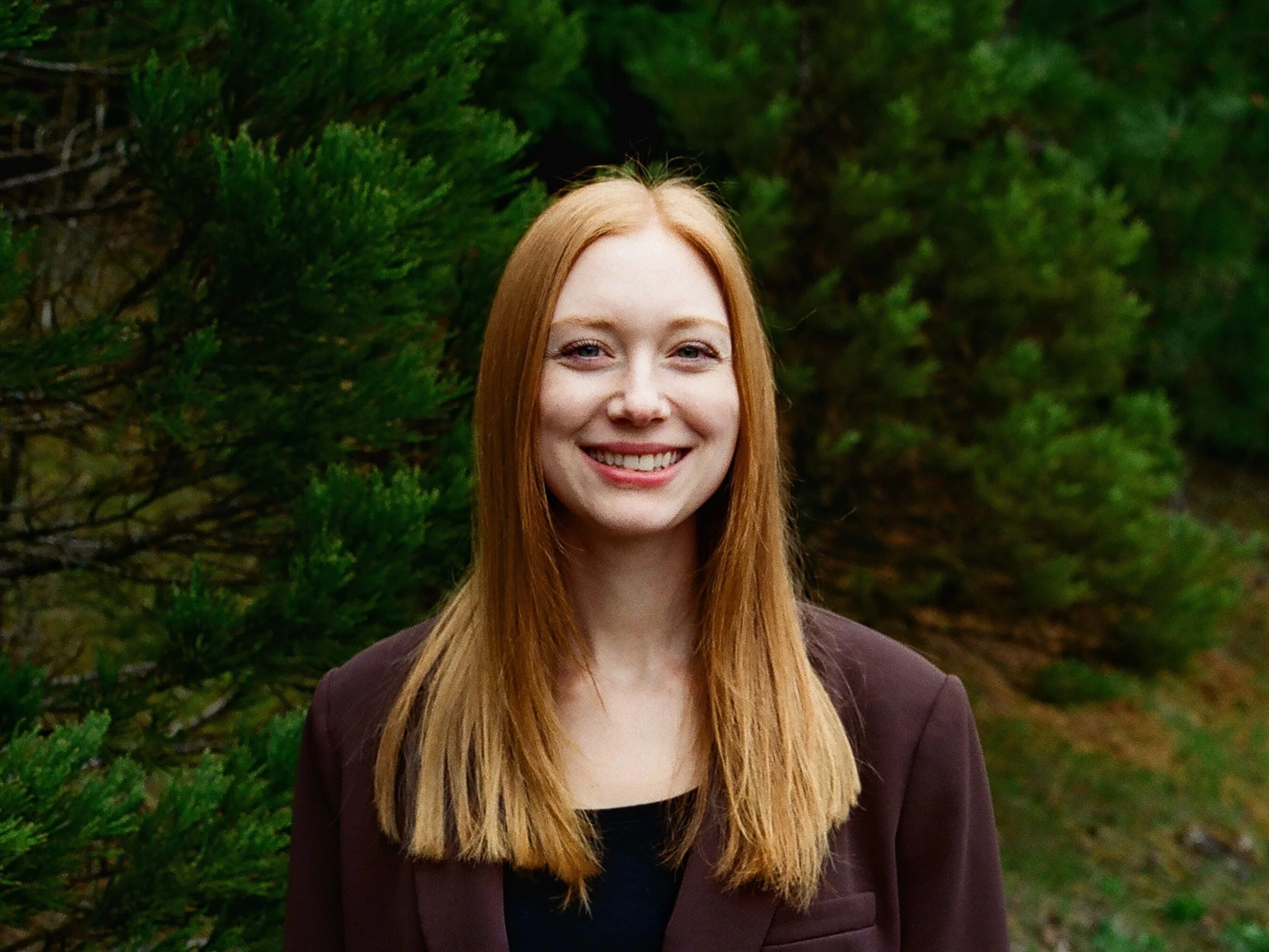 A young woman with long red hair smiling outdoors with green trees in the background.