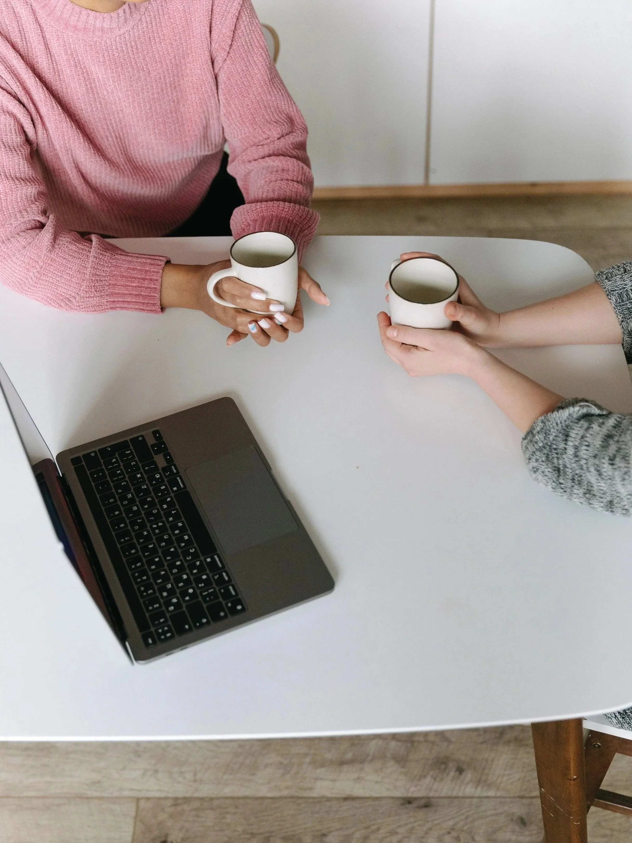 Two people sitting at a white table, each holding a white mug, with a laptop on the table and a wooden chair in the foreground.