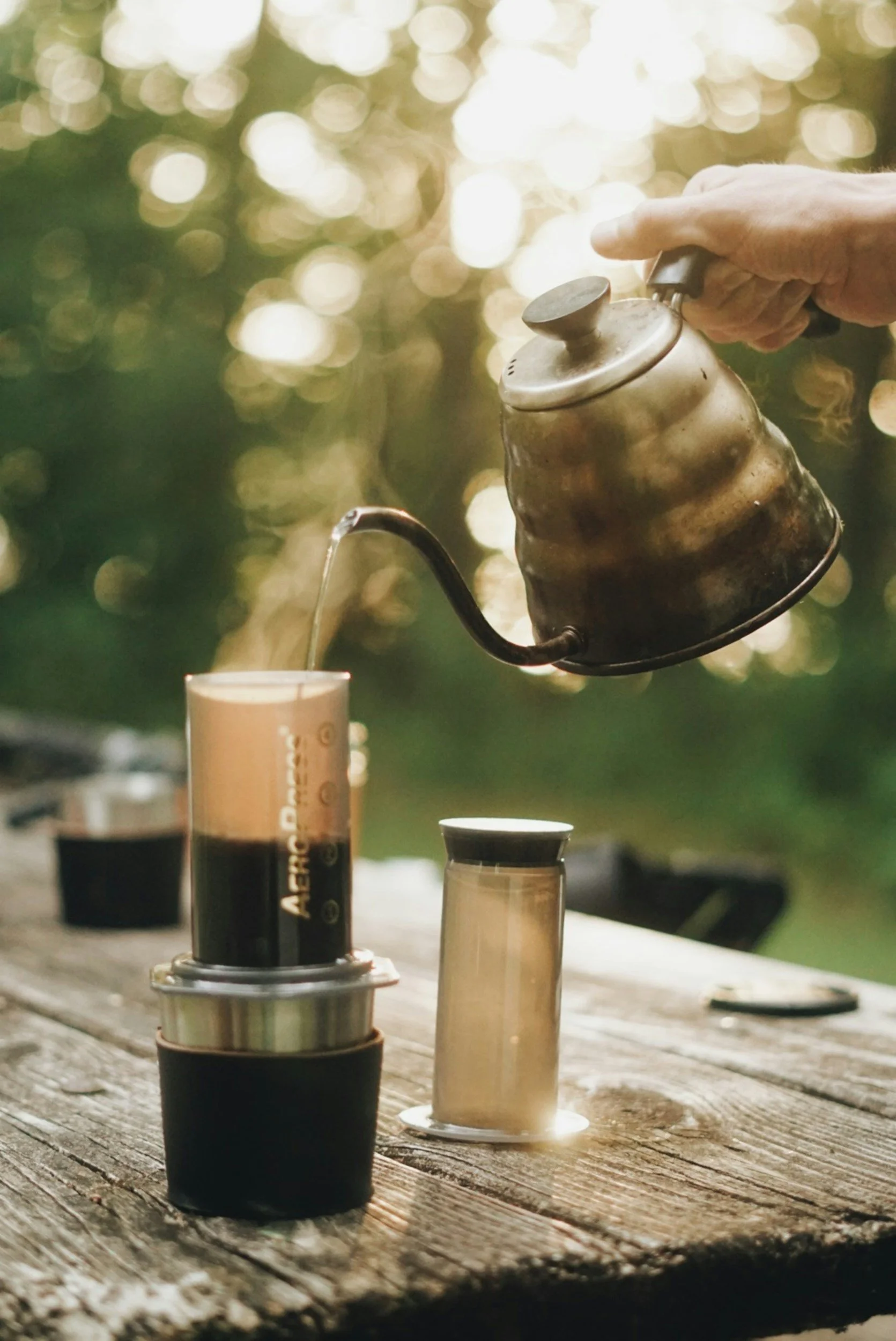 A hand pouring hot water from a vintage metal kettle into a Turkish coffee cup on a rustic wooden table outdoors with blurred greenery in the background.