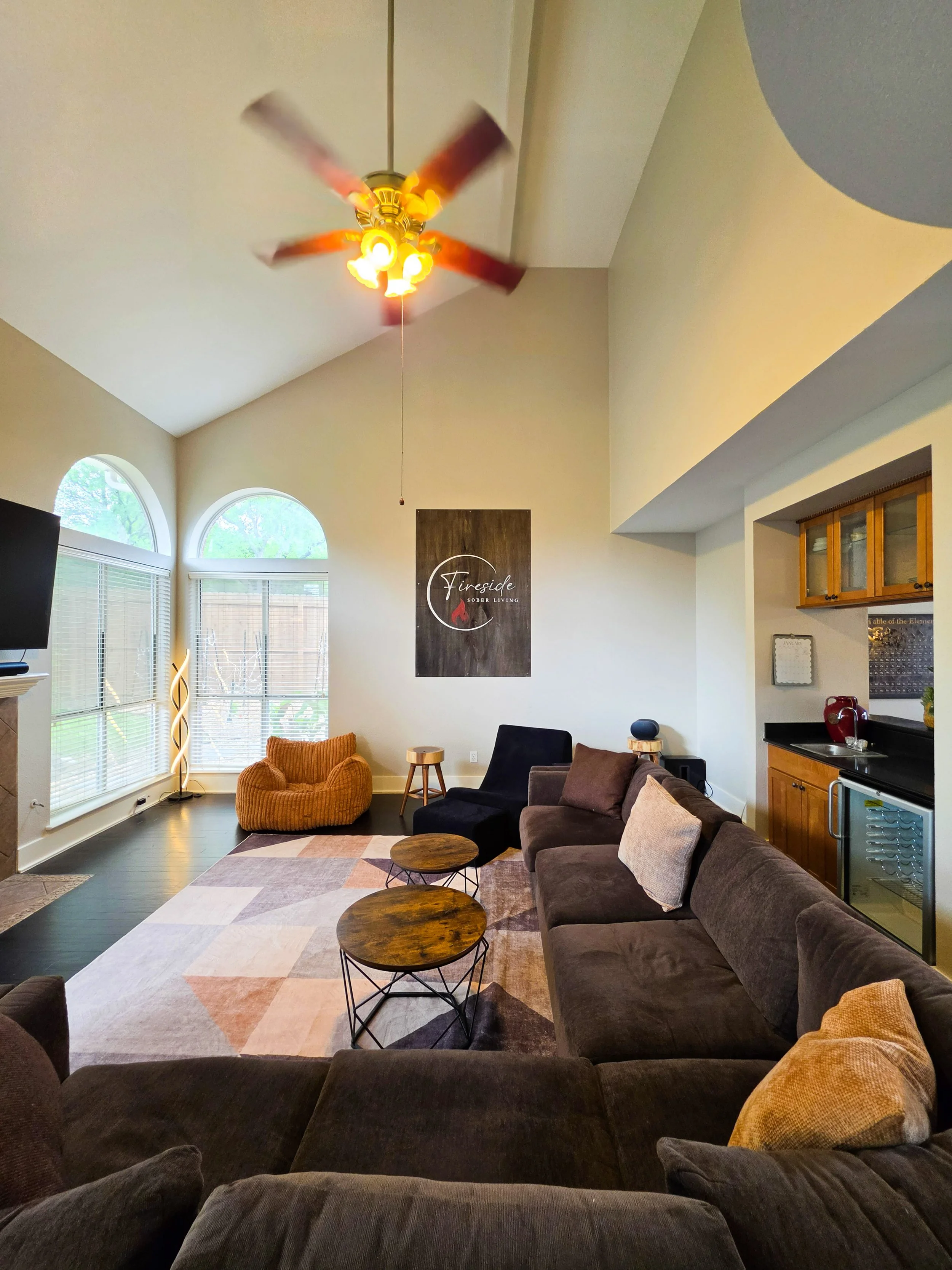 Living room with a vaulted ceiling, ceiling fan, large windows, sectional sofa, armchair, and coffee tables. Fireside Sober Living offers alternative, non-AA, non-traditional, AA, and traditional program modalities.