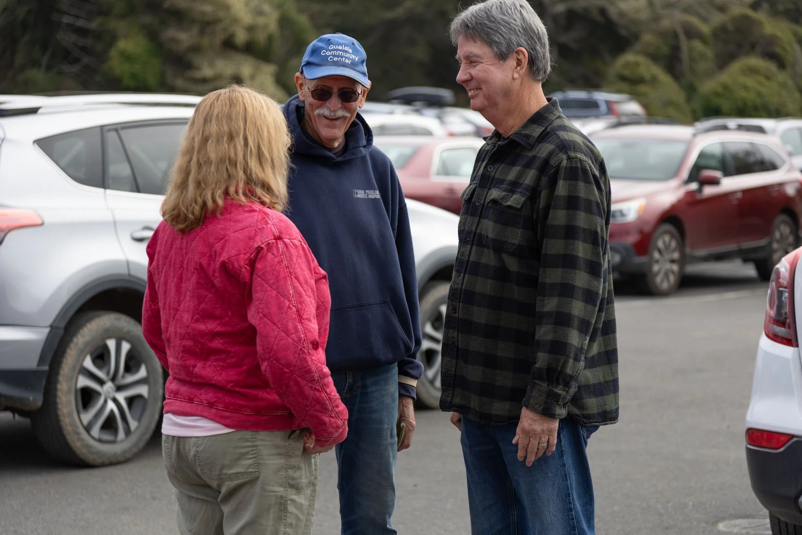 Three people standing and talking in a parking lot with several cars. Two men and one woman, all casually dressed, are smiling and engaged in conversation.