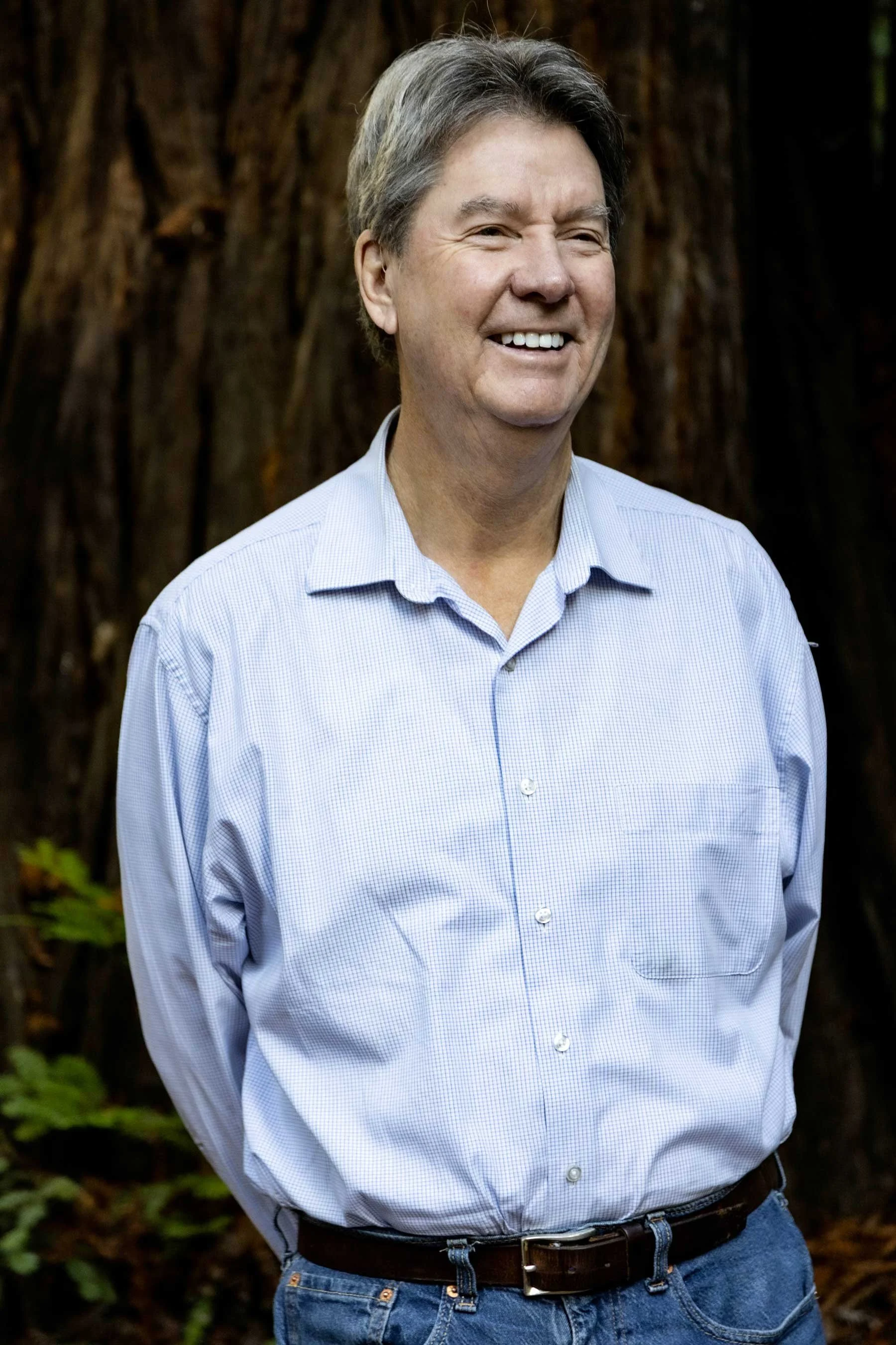 A smiling man wearing a light blue dress shirt and dark pants standing outdoors in front of a large tree trunk.